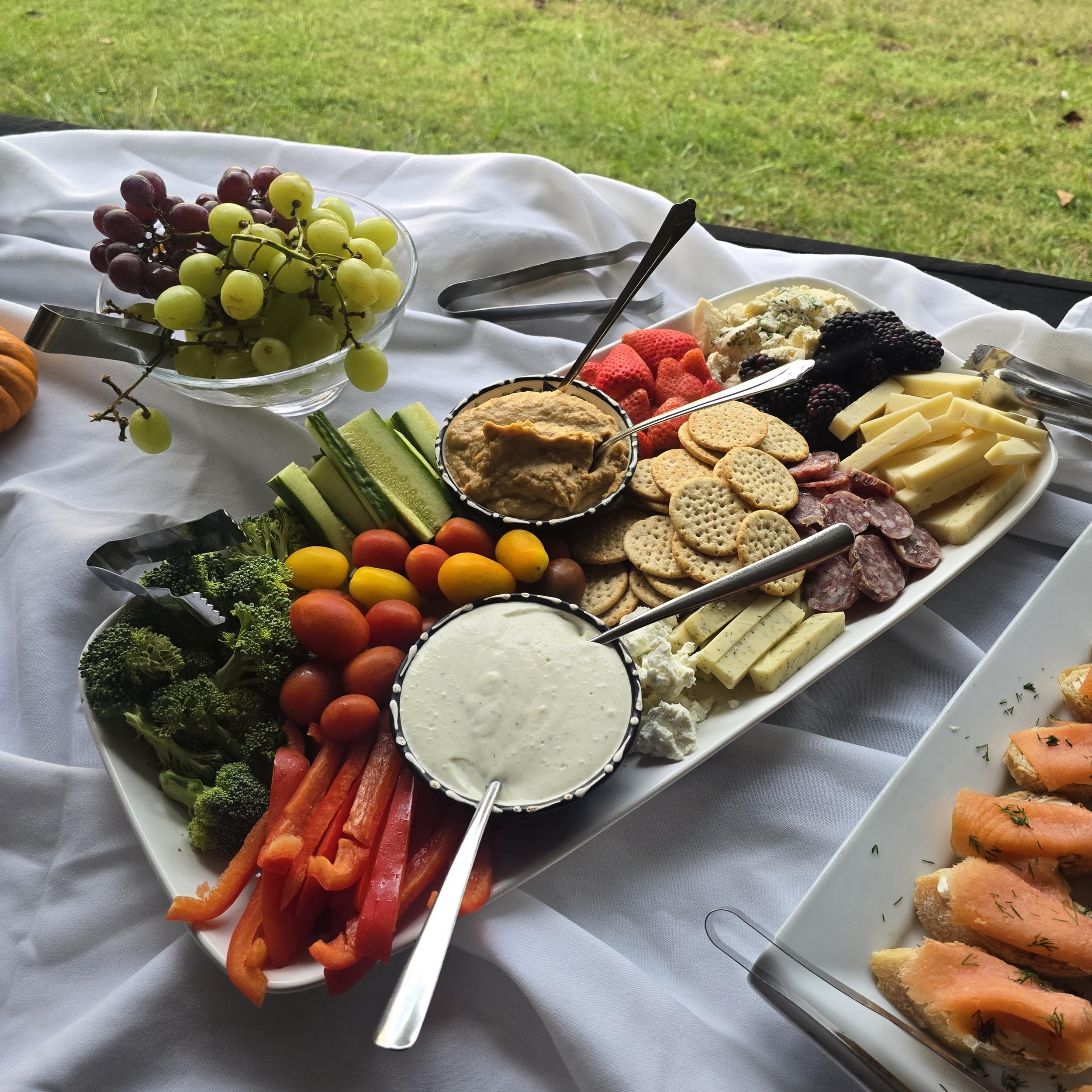 A spread of fresh vegetables, cheese, and dips on a white platter at an outdoor setting.