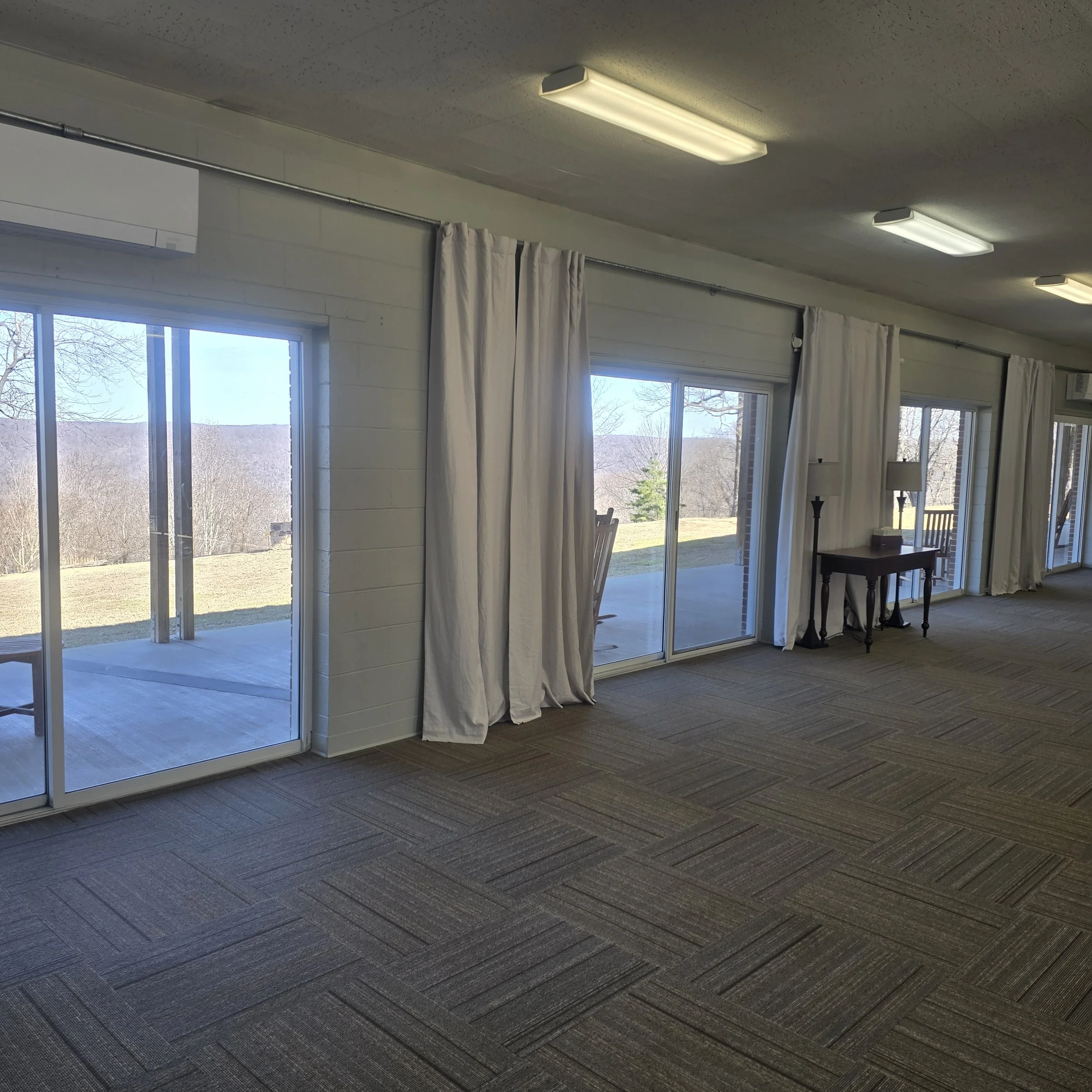 A room with large sliding glass doors leading to an outdoor patio, white curtains, a small table with two lamps, and a view of a grassy landscape with trees and hills.