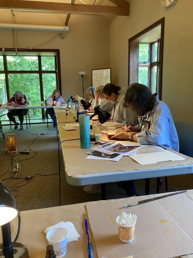 Group of women working on art projects at a long table in a well-lit room with large windows and green trees visible outside.