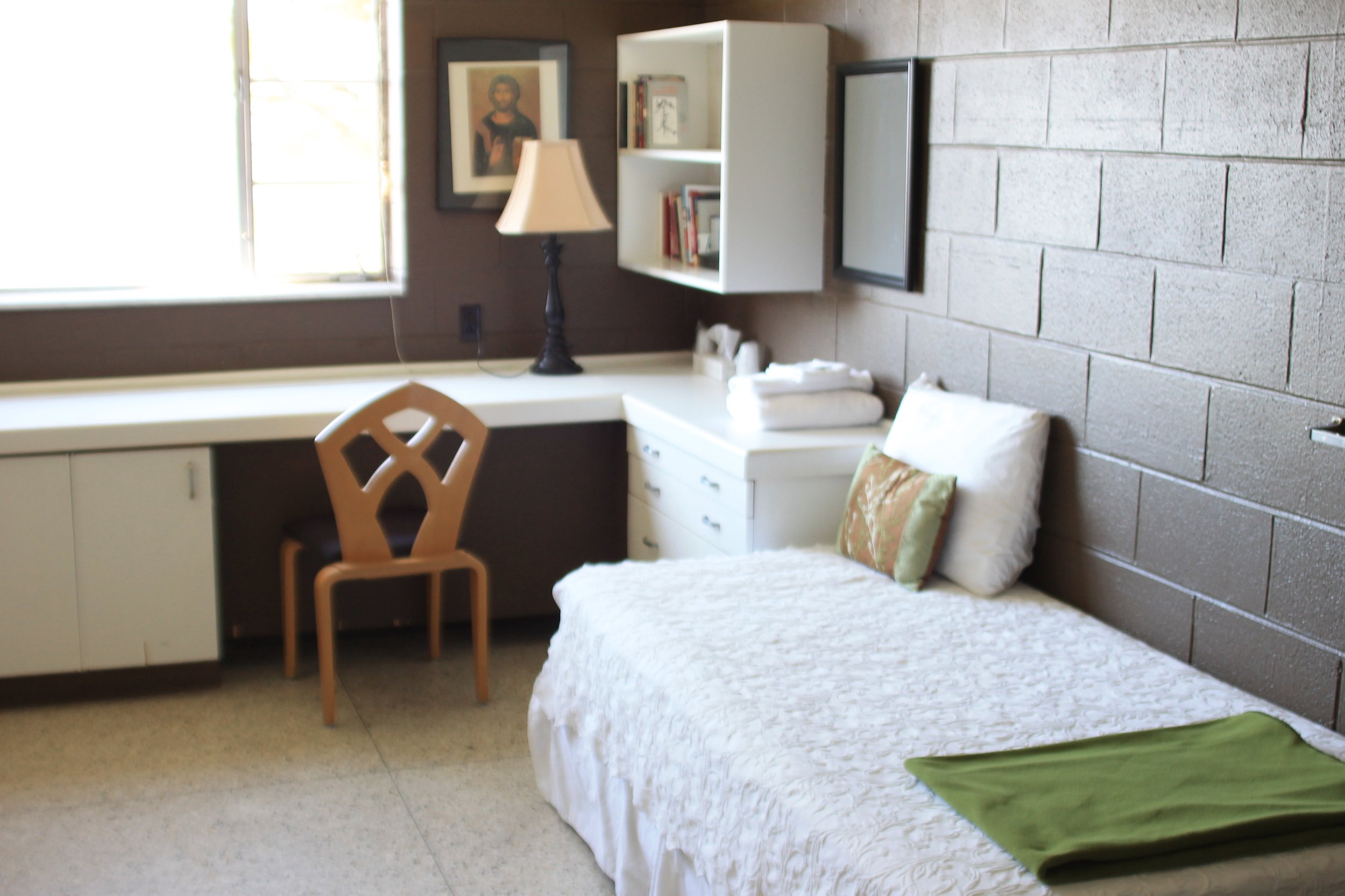 A small, simple bedroom with a single bed covered with white bedding and a green pillow. Next to the bed is a white bedside table with folded white towels. There is a wooden chair at a white desk under a window, with sunlight coming in. On the wall, 