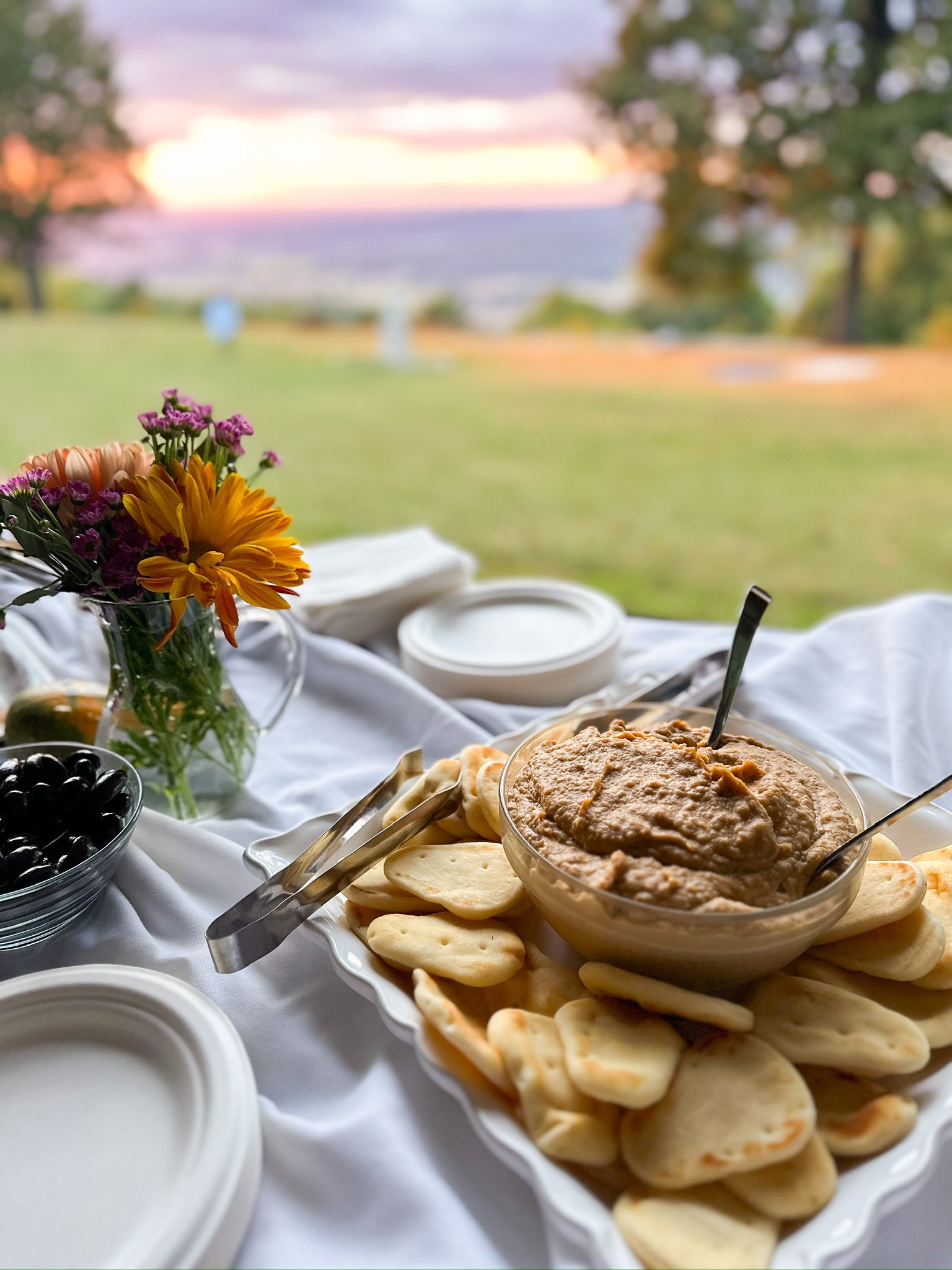 A breakfast table with a bowl of peanut butter, crackers, a bowl of black olives, a flower arrangement, and paper plates, set outdoors with a view of a sunset over a field and trees in the background.