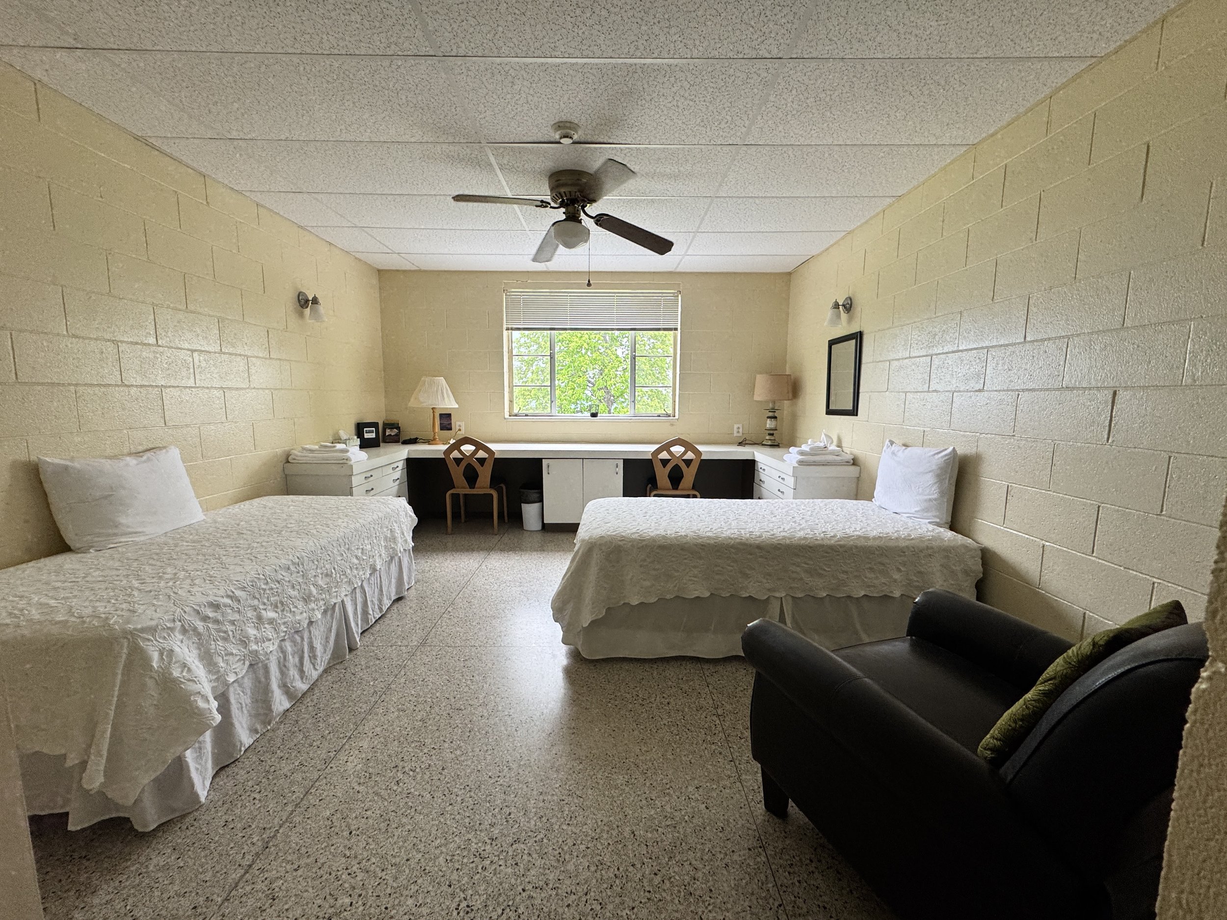 A simple room with two single beds covered in white bedding, a black armchair, a window with a view of green trees, desk area with two chairs and lamps, and beige brick walls.