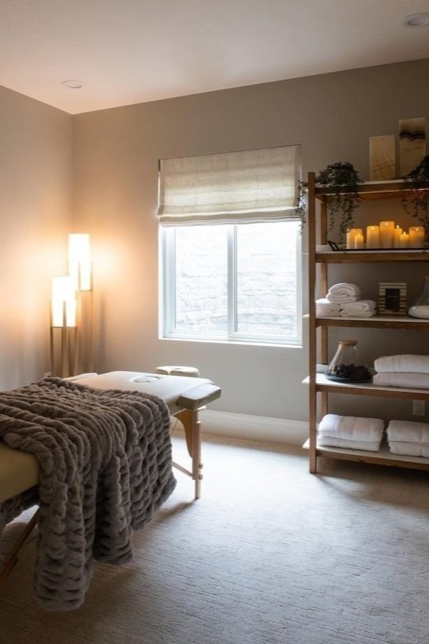 A cozy massage therapy room with a massage table draped in a soft, furry blanket next to a window. There is a wooden shelf holding candles, rolled towels, and decorative items, creating a relaxing atmosphere.