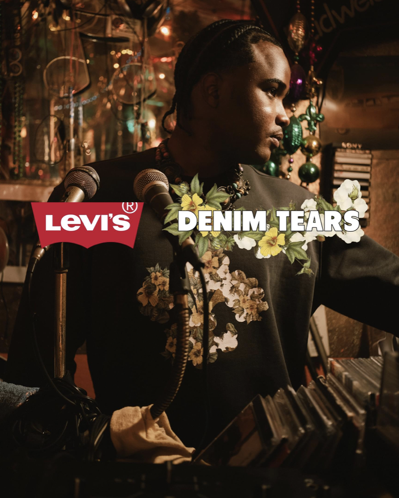 A person with braided hair and floral necklace, standing in a dimly lit room decorated with Christmas ornaments, with microphones in front, representing Levi's Denim Tears.