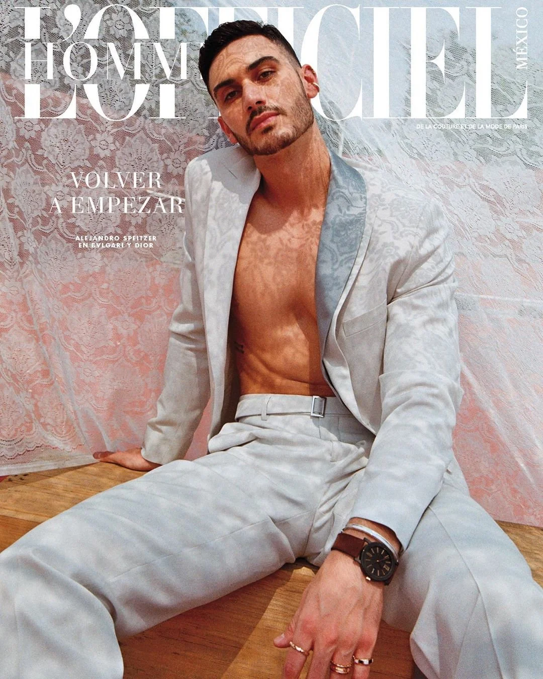 A man in a light-colored suit with no shirt, sitting on a wooden floor against a pink and lace-patterned background, with large magazine text overlay reading 'L'Officiel Homme Mexico' and other French text.
