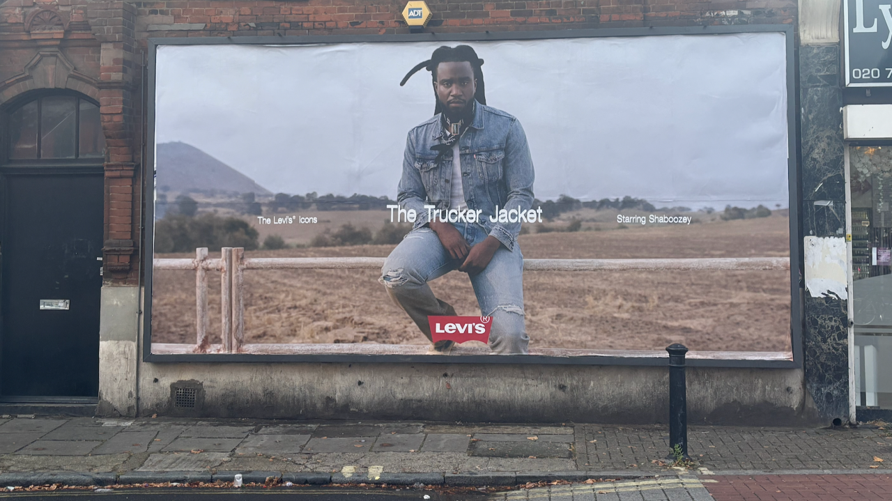 Levi's advertisement featuring a man with dreadlocks sitting on a fence in an open field, wearing a denim jacket and jeans, with mountains in the background.