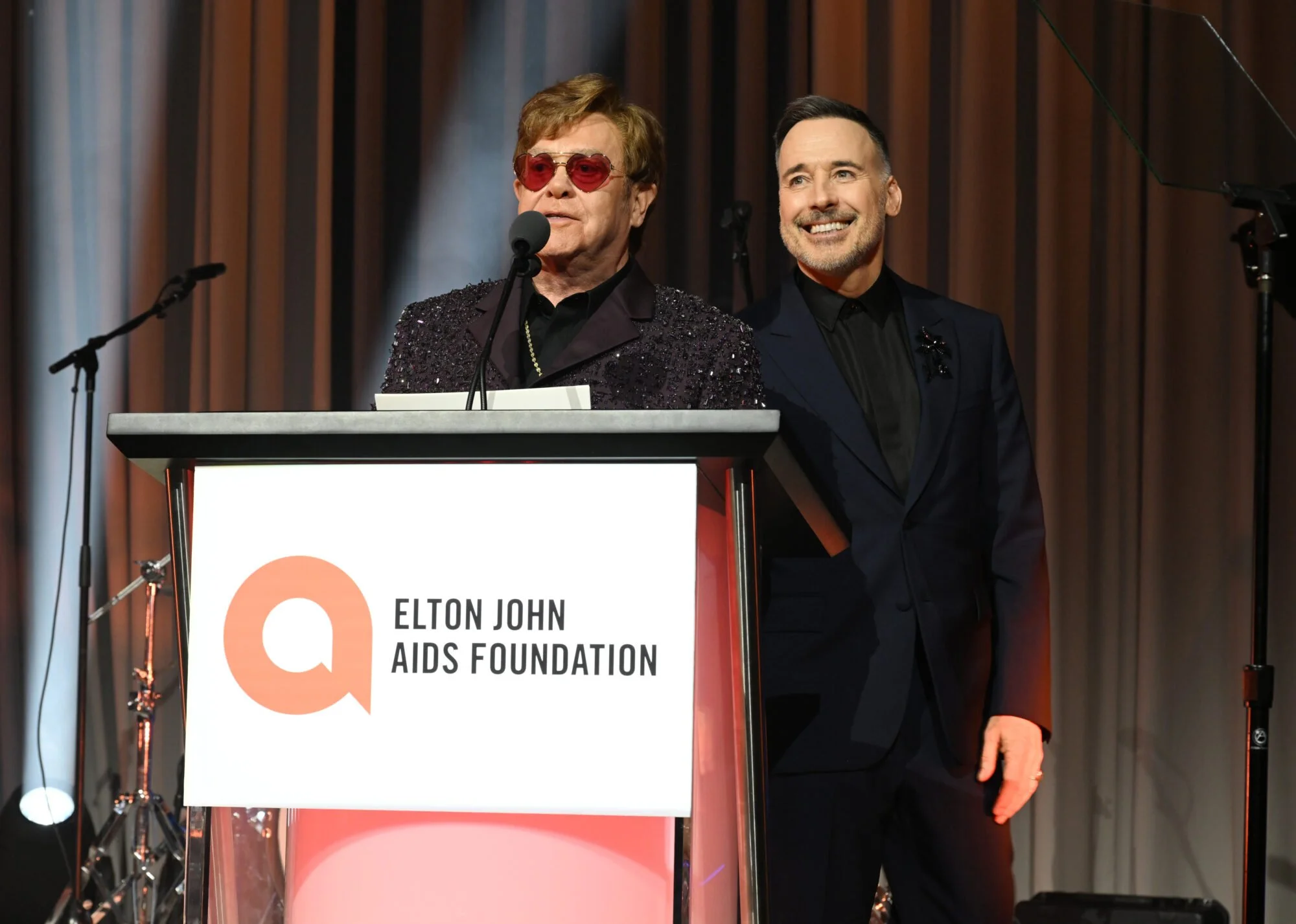 Elton John speaking at a podium during an event for the Elton John AIDS Foundation, with a man standing beside him on stage.