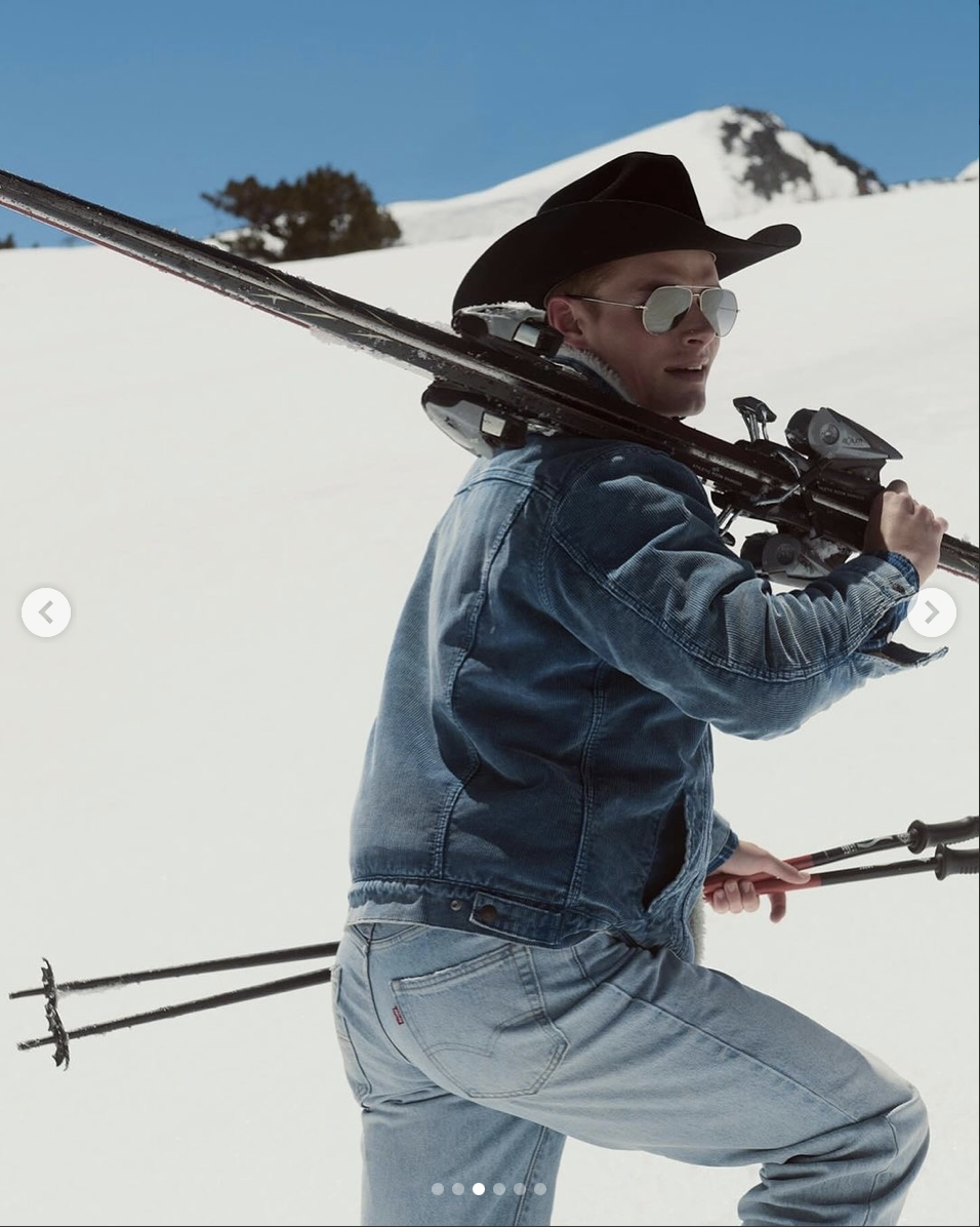 A man in a black cowboy hat, sunglasses, a denim jacket, and light jeans is carrying skis over his shoulder while walking in a snowy mountain landscape with a clear blue sky.