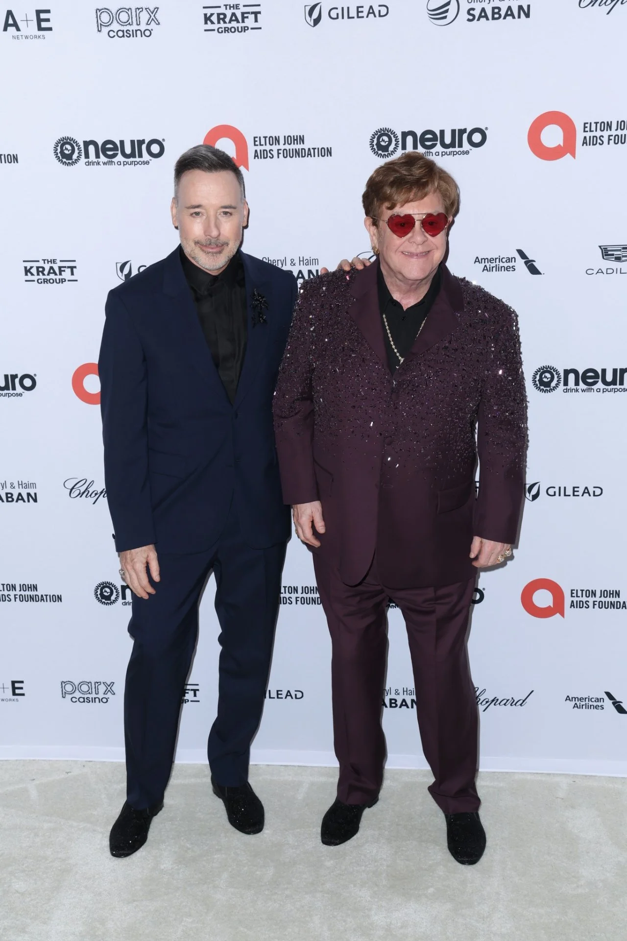 Two men in tuxedos standing together on a red carpet. The man on the left has short hair and a beard, wearing a dark blue tuxedo, black shirt, and black shoes. The man on the right has short hair, wearing red sunglasses, a purple sequin blazer, black