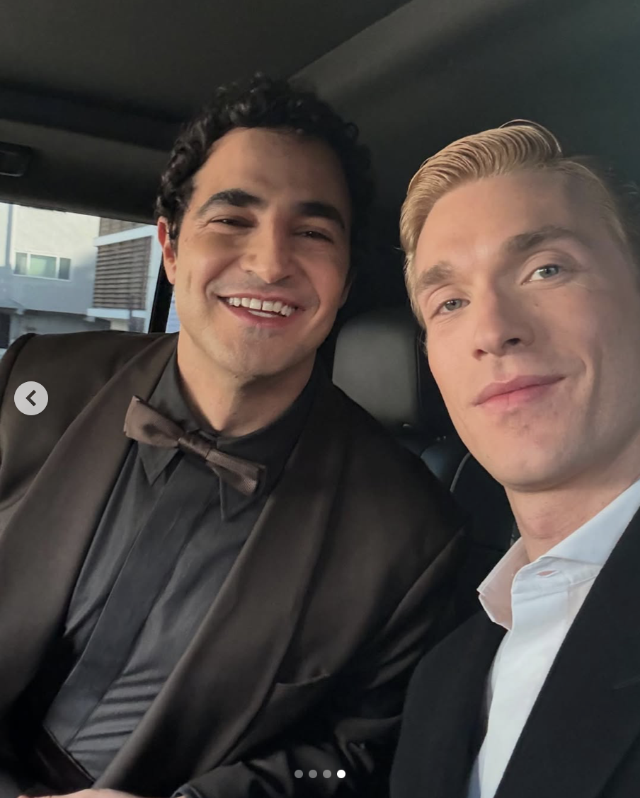 Two young men in formal attire, smiling, seated inside a vehicle, with the city skyline visible through the windows.