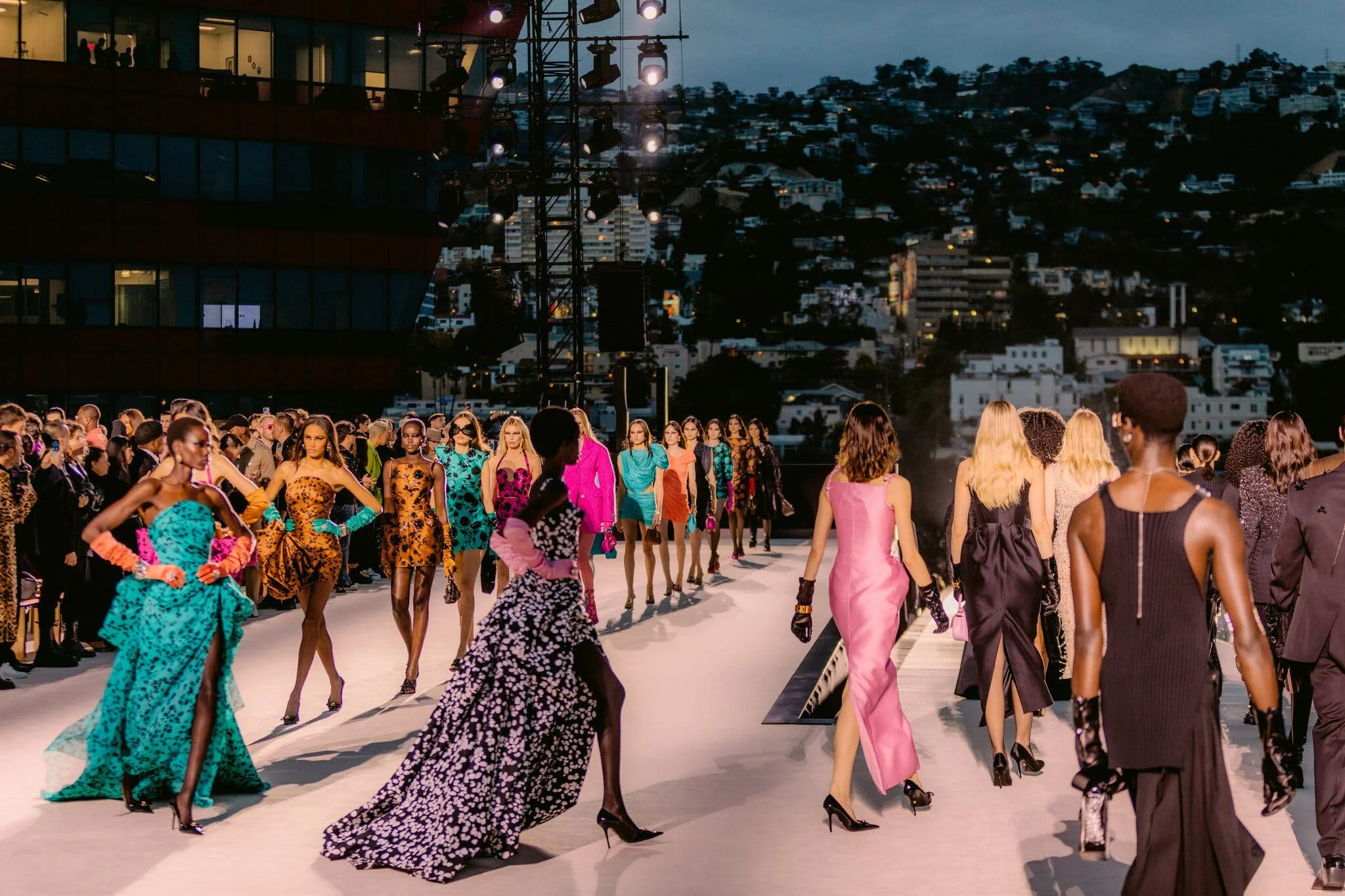 Fashion models walk runway during a fashion show at dusk, with city buildings and a hill in the background.
