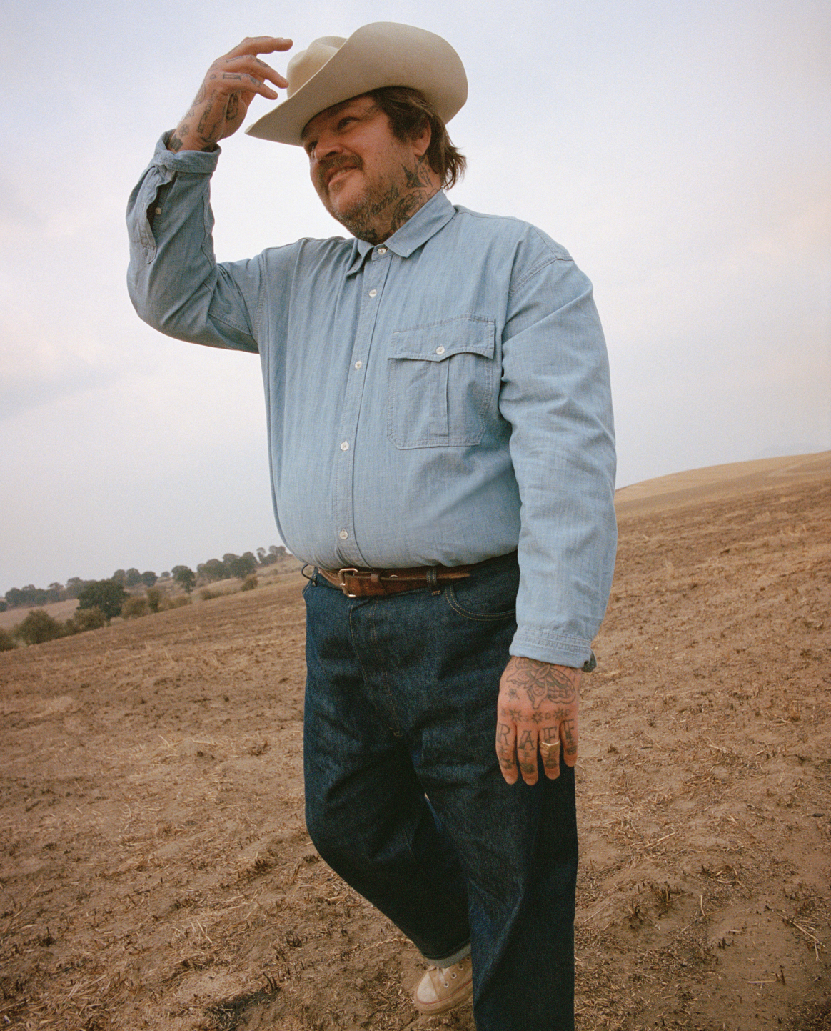 A man with tattoos and a beard, wearing a cowboy hat, denim shirt, and jeans, standing in a dry, open field.