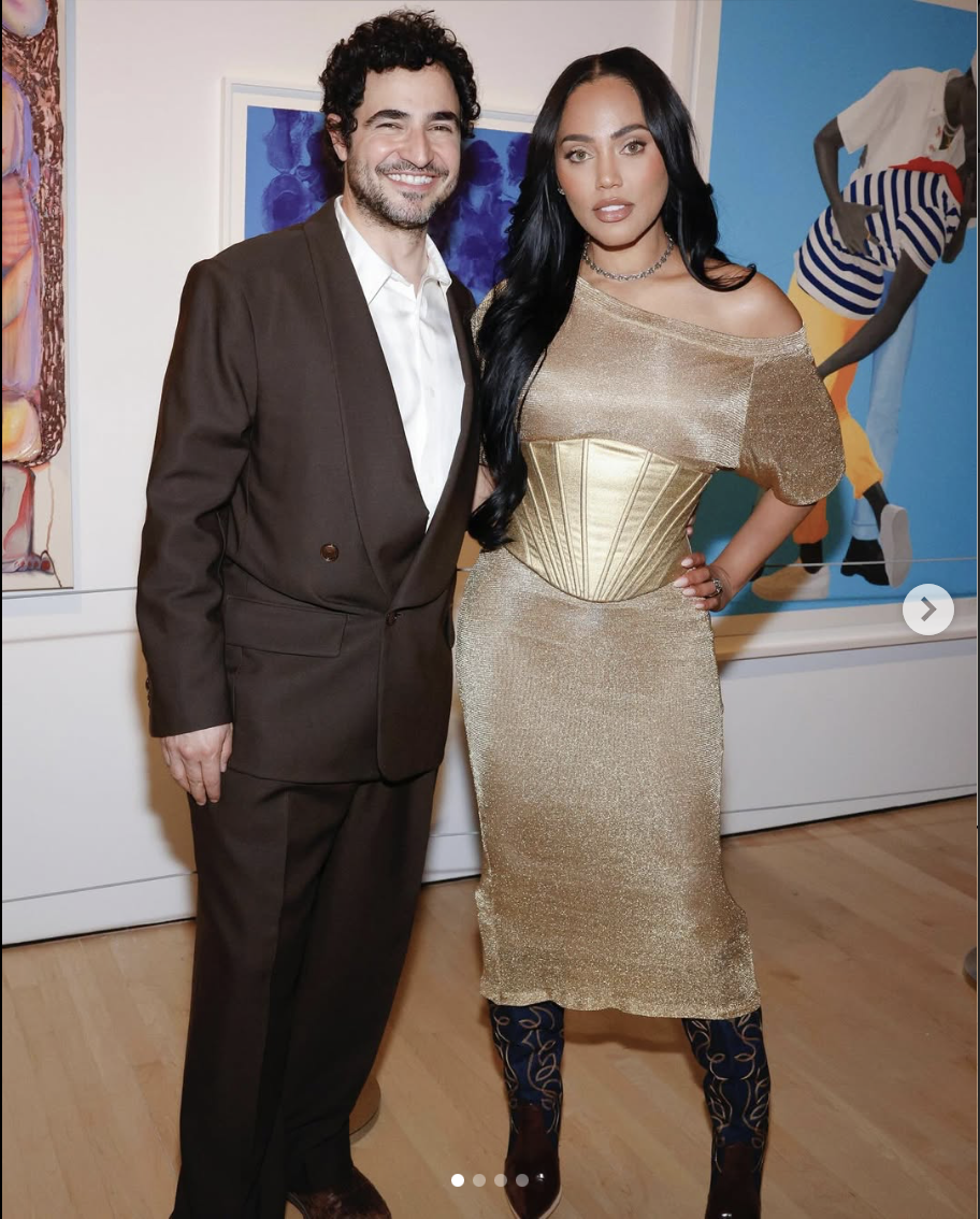 A man and a woman standing together at an art gallery. The man is wearing a brown suit with a white shirt underneath, and the woman is wearing a shimmery gold dress with a corset-style waist, and patterned black and gold tights.