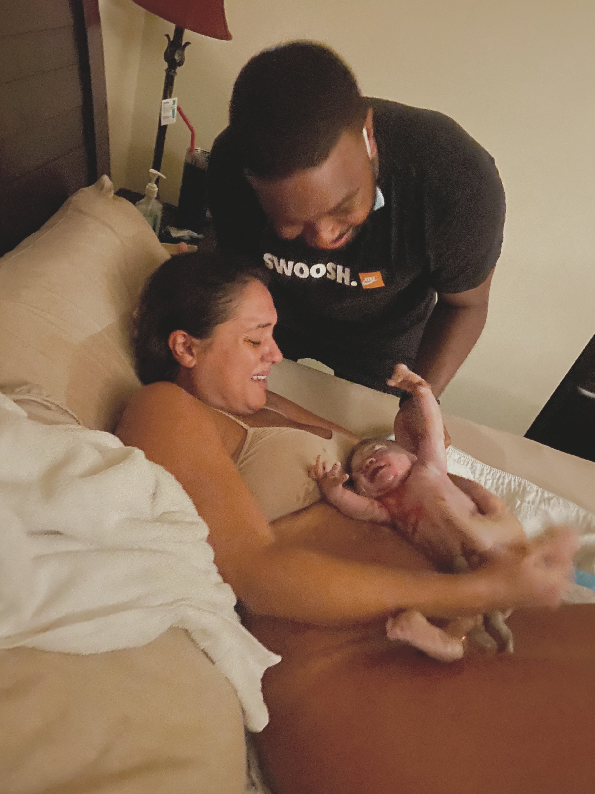 A woman in labor holding a newborn baby in a hospital bed while a man stands over her smiling.