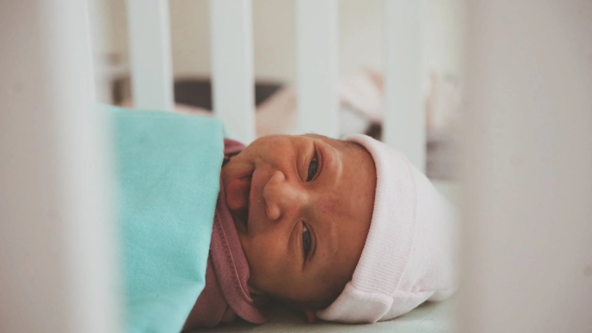 Newborn baby lying in a hospital crib, wearing a white hat and pink outfit.