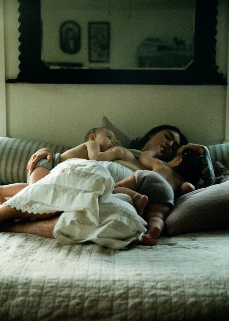A woman and two young children cuddling on a bed, lying close to each other. The woman is lying on her side with a child resting on her, and another child is lying across her and the first child.