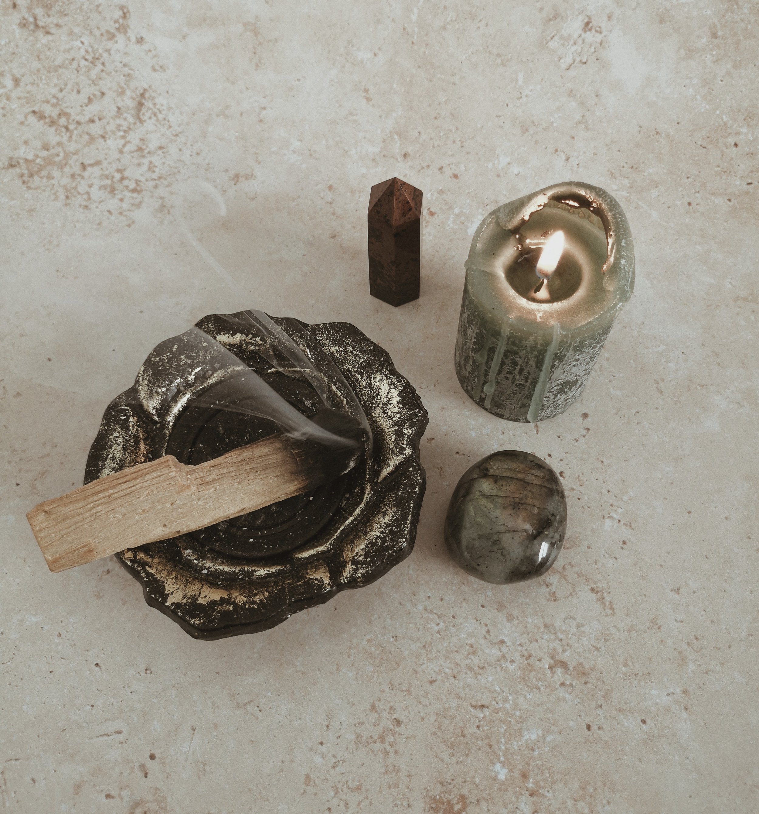 A collection of objects on a light-colored surface, including a black stone incense holder with a stick of incense, a lit green candle, a small rectangular stone, and a polished stone orb.