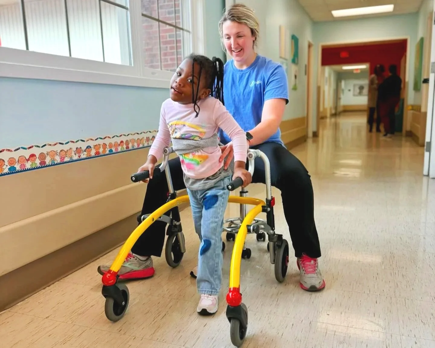 A young girl walking with a walker, assisted by a smiling woman in a blue shirt, in a bright hallway of a healthcare facility.
