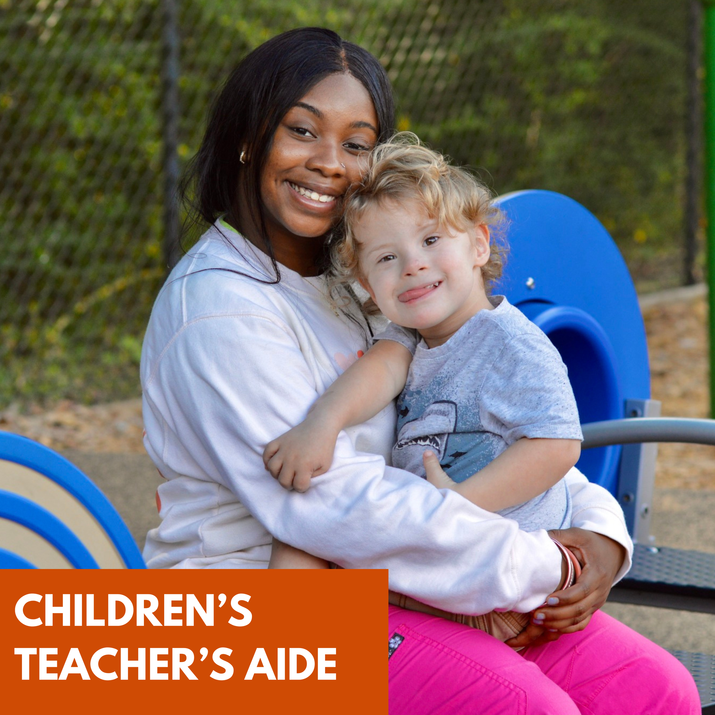 A young woman with dark hair and a white sweatshirt holds and smiles at a smiling young boy with curly blond hair, wearing a gray t-shirt. They are outdoors on a playground, with trees and a fence in the background.