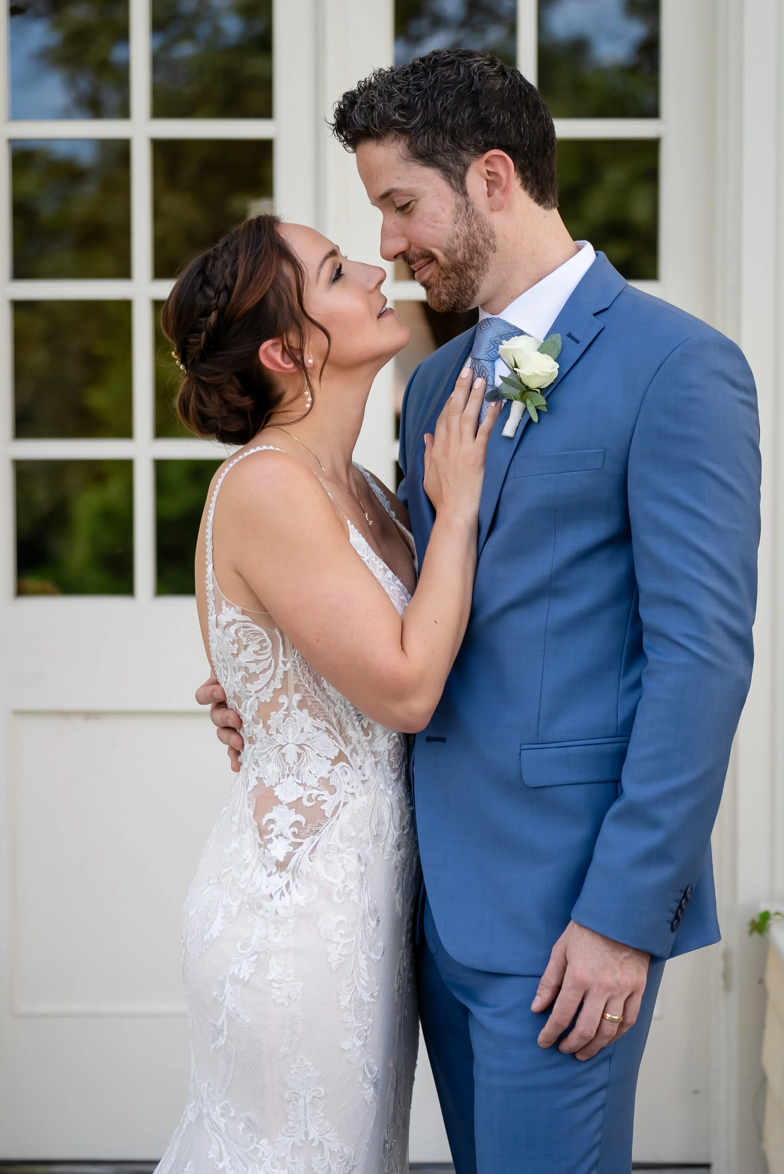 A bride and groom standing close together indoors by a white door with windows, gazing into each other's eyes, with the groom touching the bride's waist and the bride holding his chest.