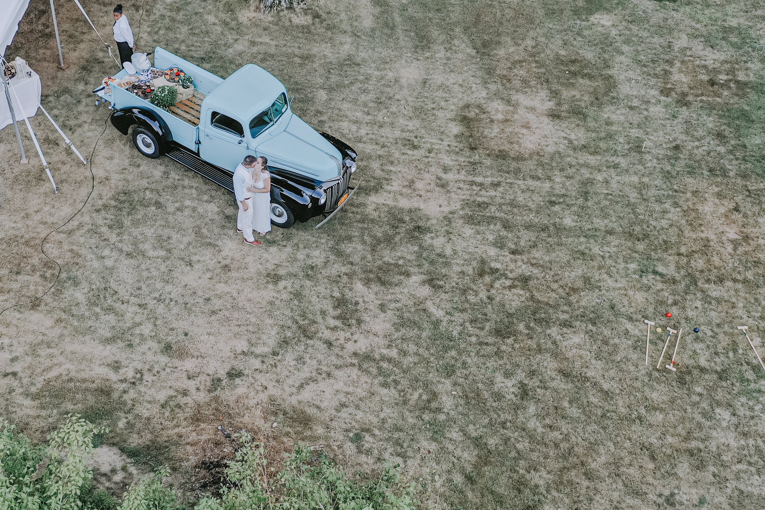 A light blue vintage pickup truck with black fenders and a black front, parked on a grassy field. Two people, a man and a woman, are standing close together near the front of the truck, possibly posing for a photo. The truck has a flatbed with some g