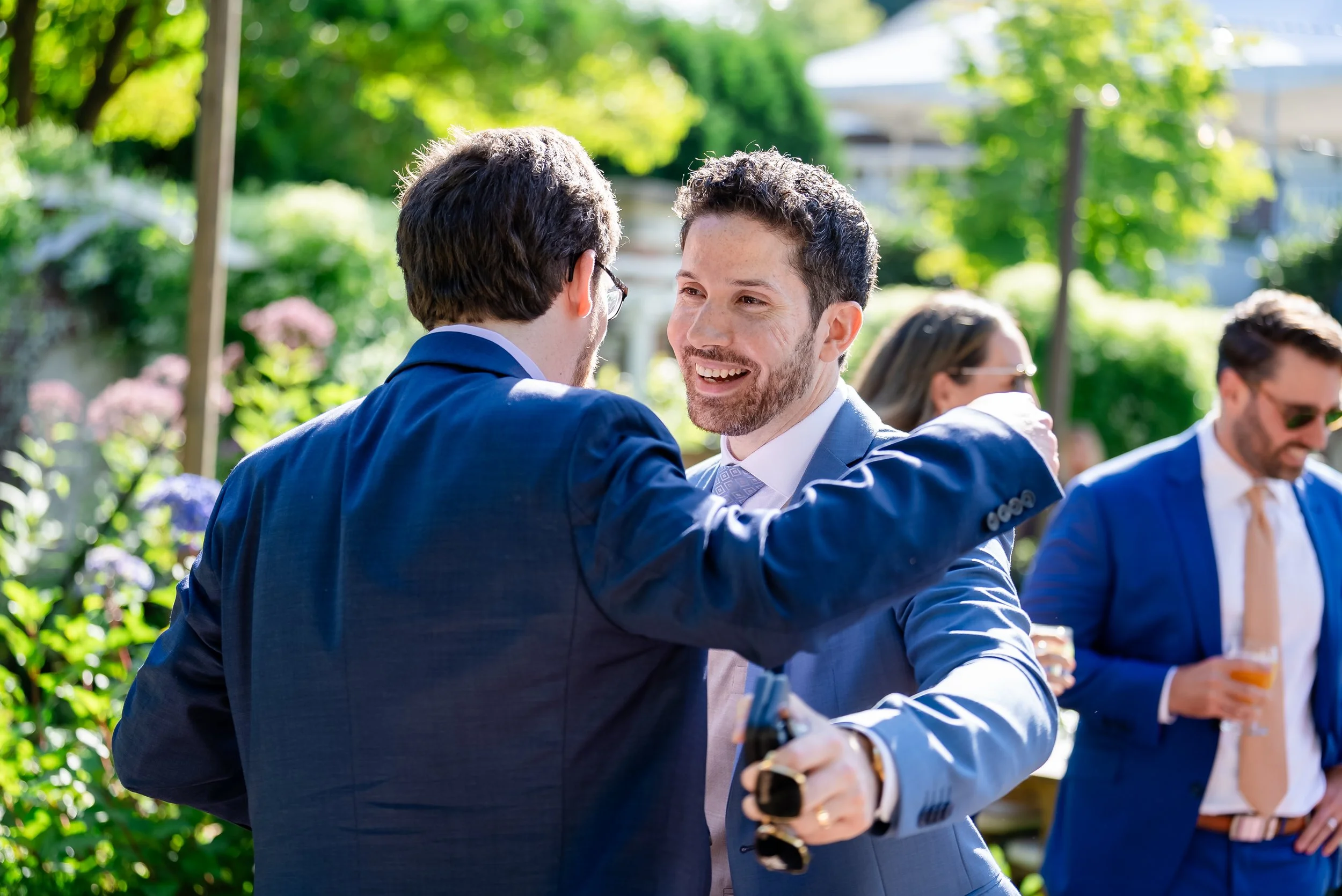 Two men in suits smiling and greeting each other outdoors at a social event with trees and other people in the background.