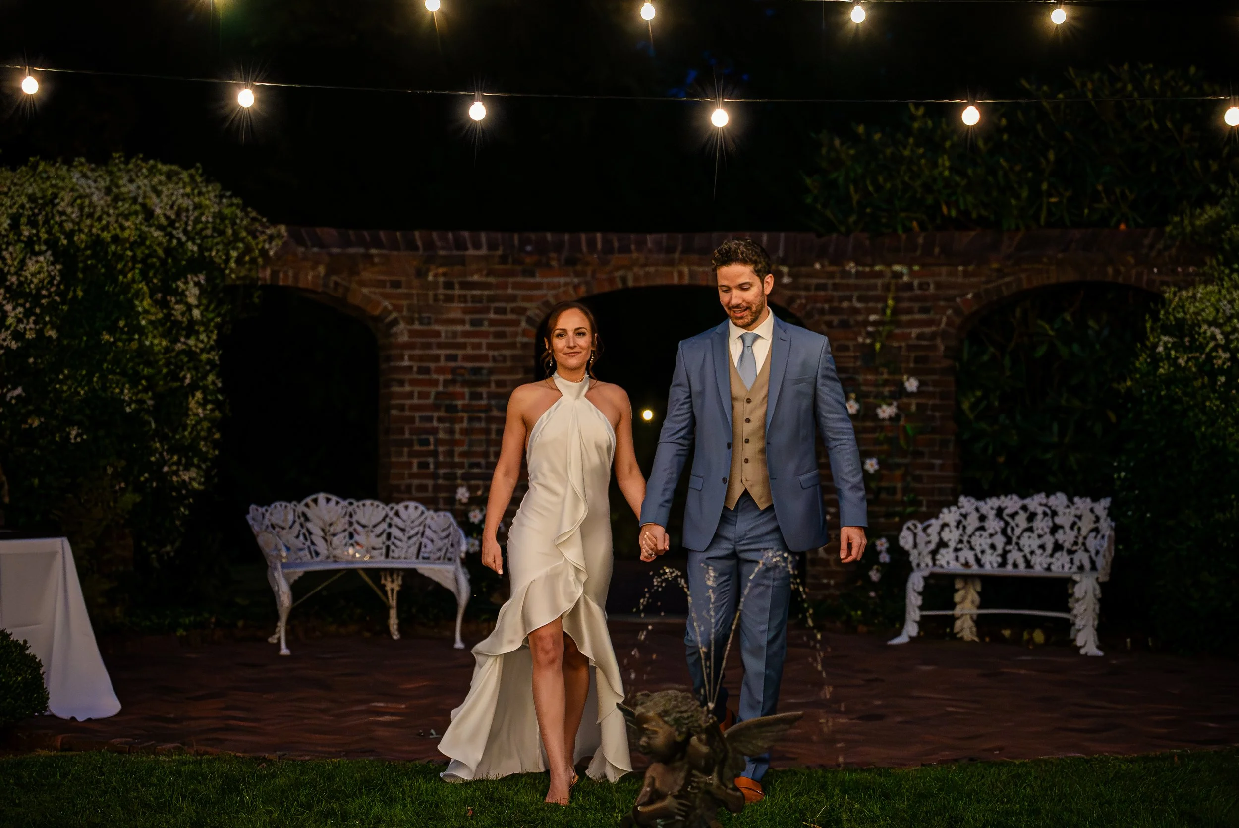 A bride and groom walking hand-in-hand outdoors at night during their wedding reception, illuminated by hanging string lights and surrounded by decorated chairs and greenery.