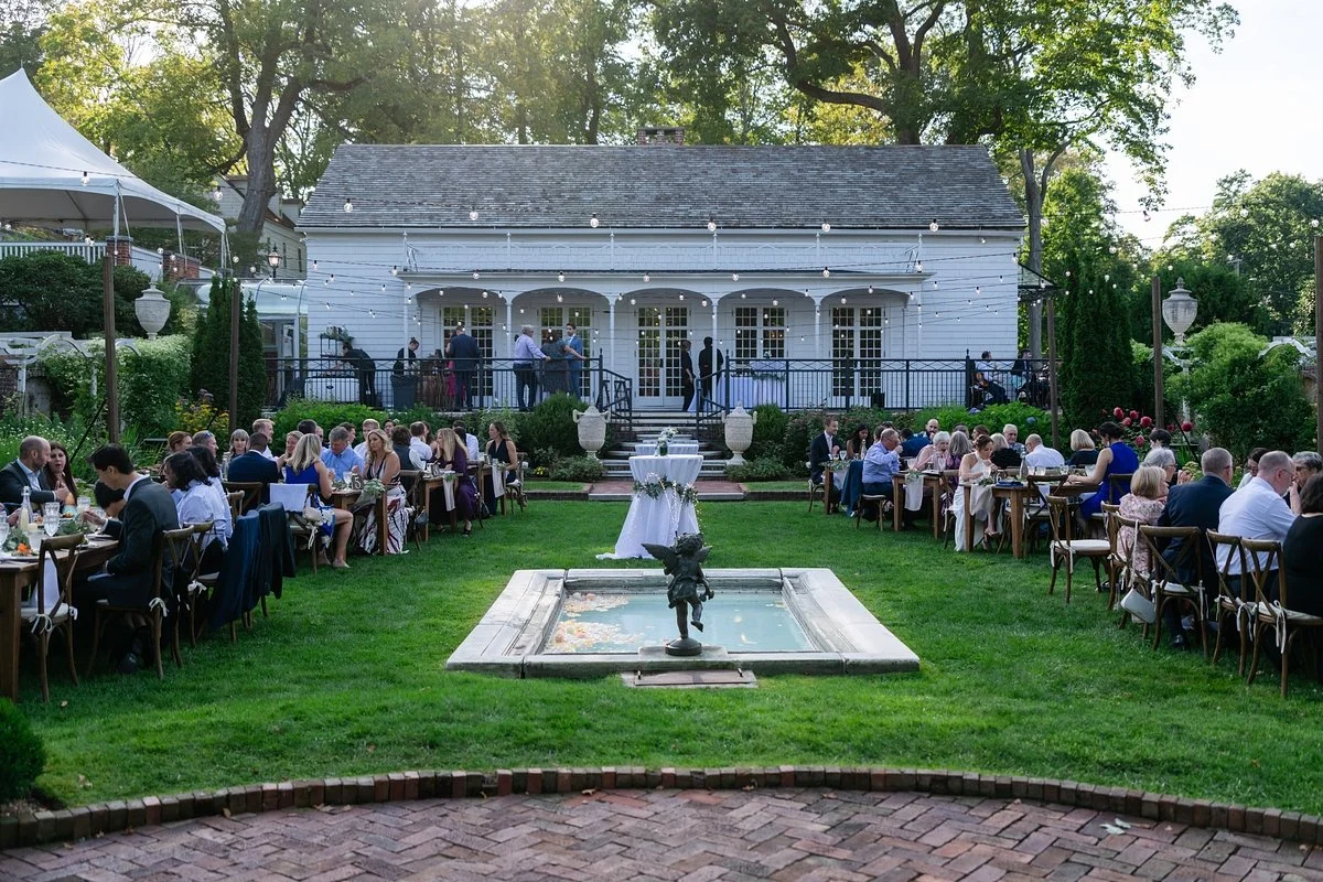 Elegant outdoor wedding reception in a lush garden with a fountain, long tables with guests, and a white house in the background decorated with string lights in the evening.