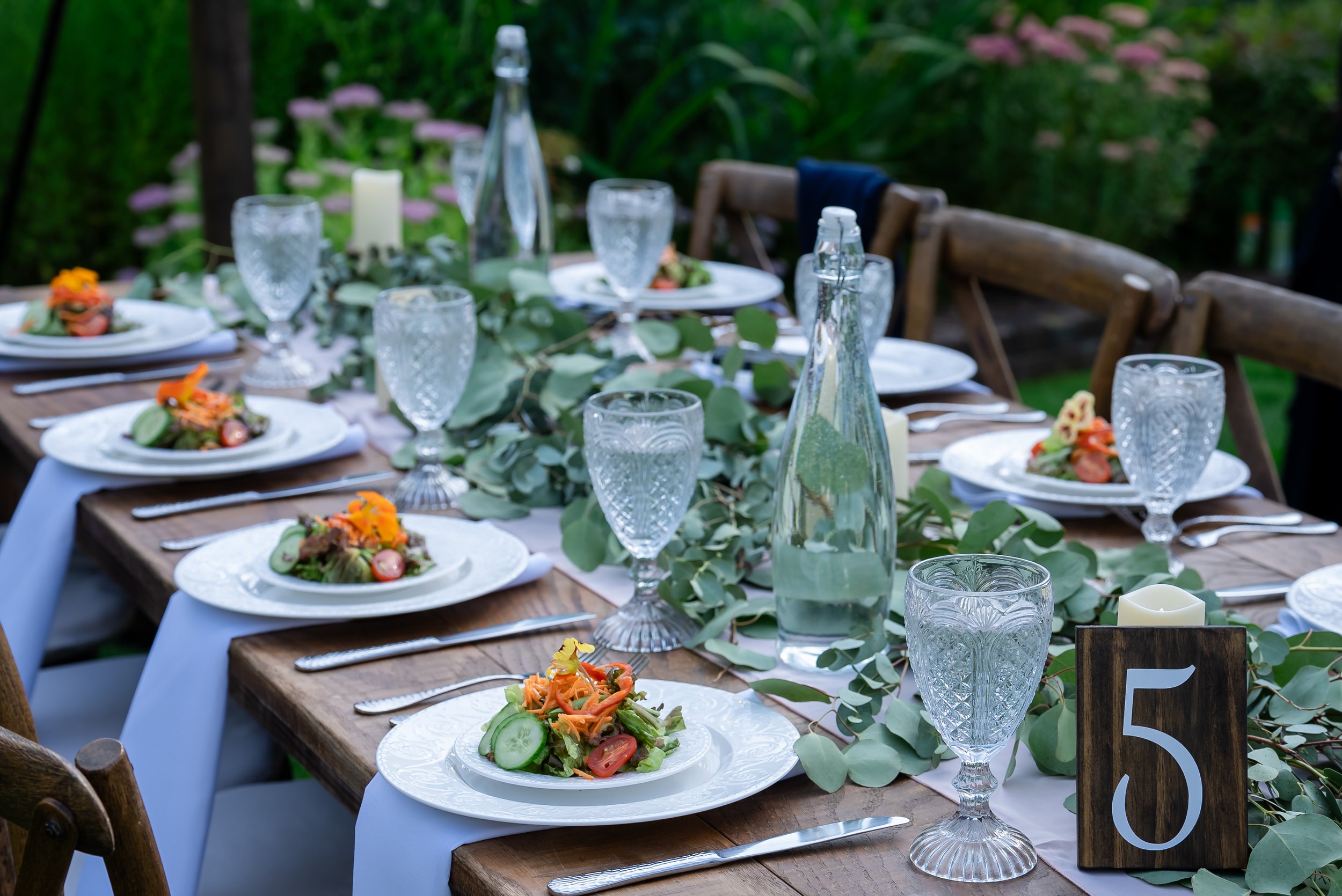 Outdoor dining table set for a celebration with salad plates, water glasses, bottles, and candles, decorated with green foliage and flowers in a garden.