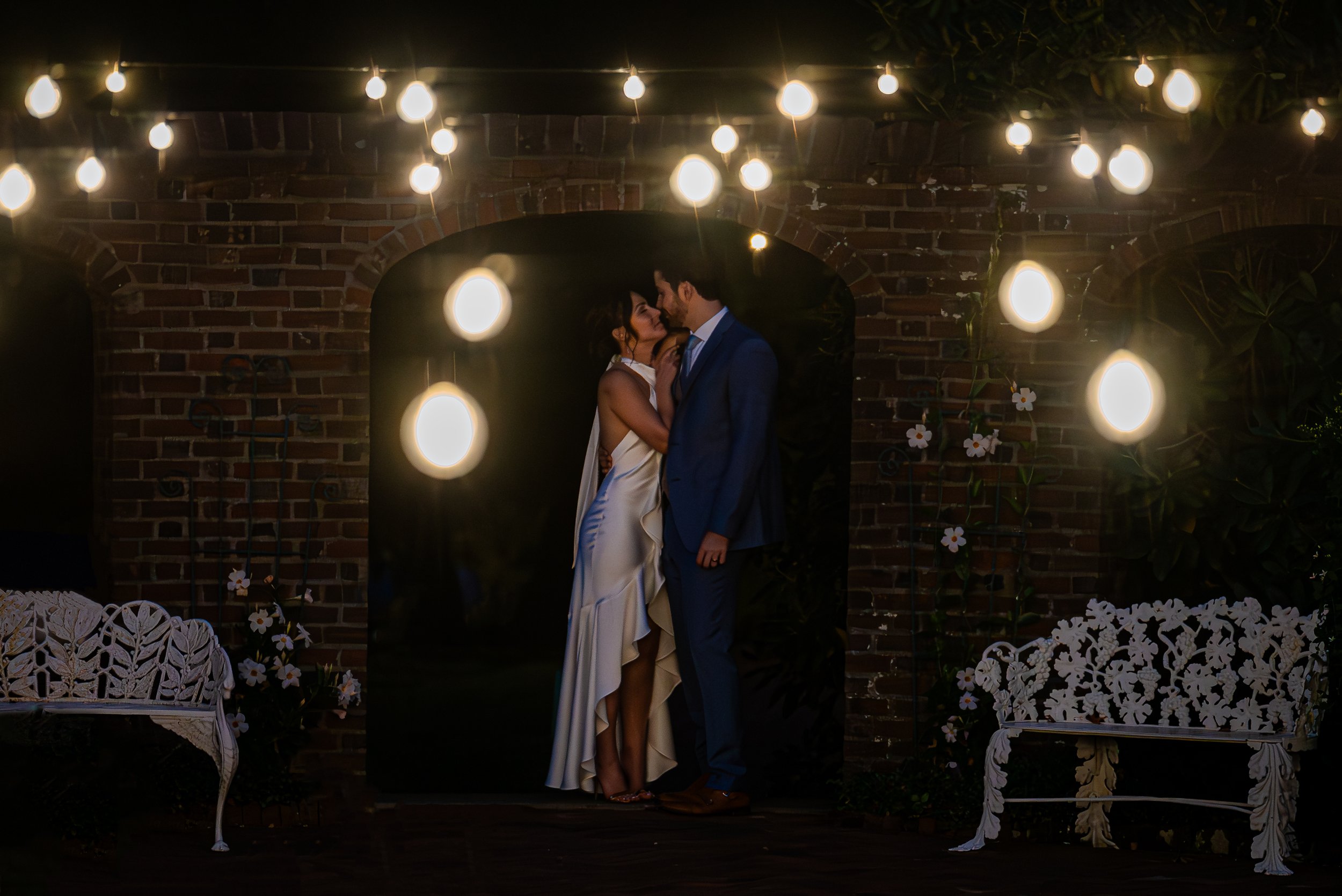 A couple in wedding attire sharing an intimate moment under hanging string lights during the night, with a brick wall and garden decorations around them.