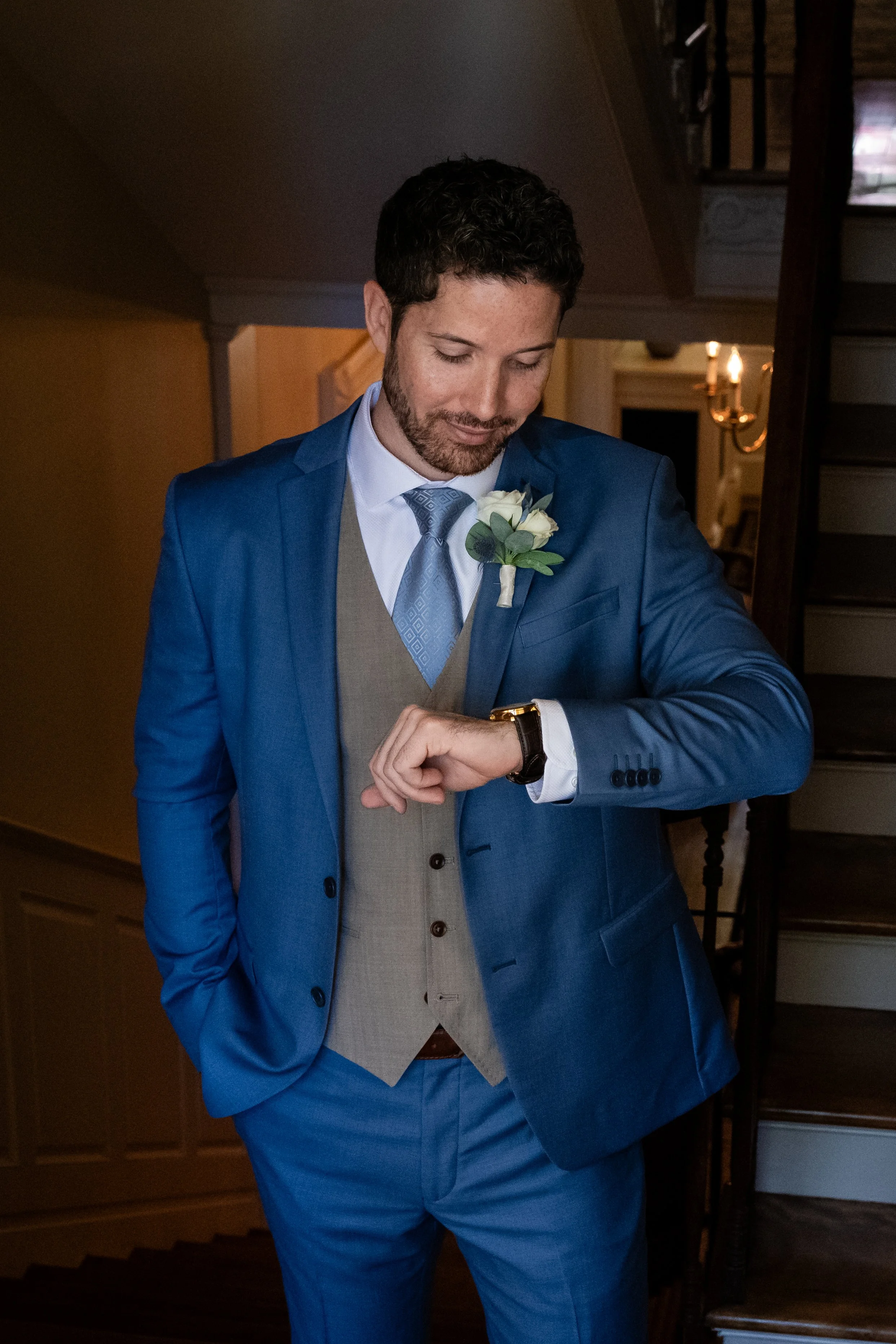 A man in a blue suit, white shirt, and blue patterned tie, with a white rose boutonnière, looking at his watch inside a warmly lit house, near a staircase.