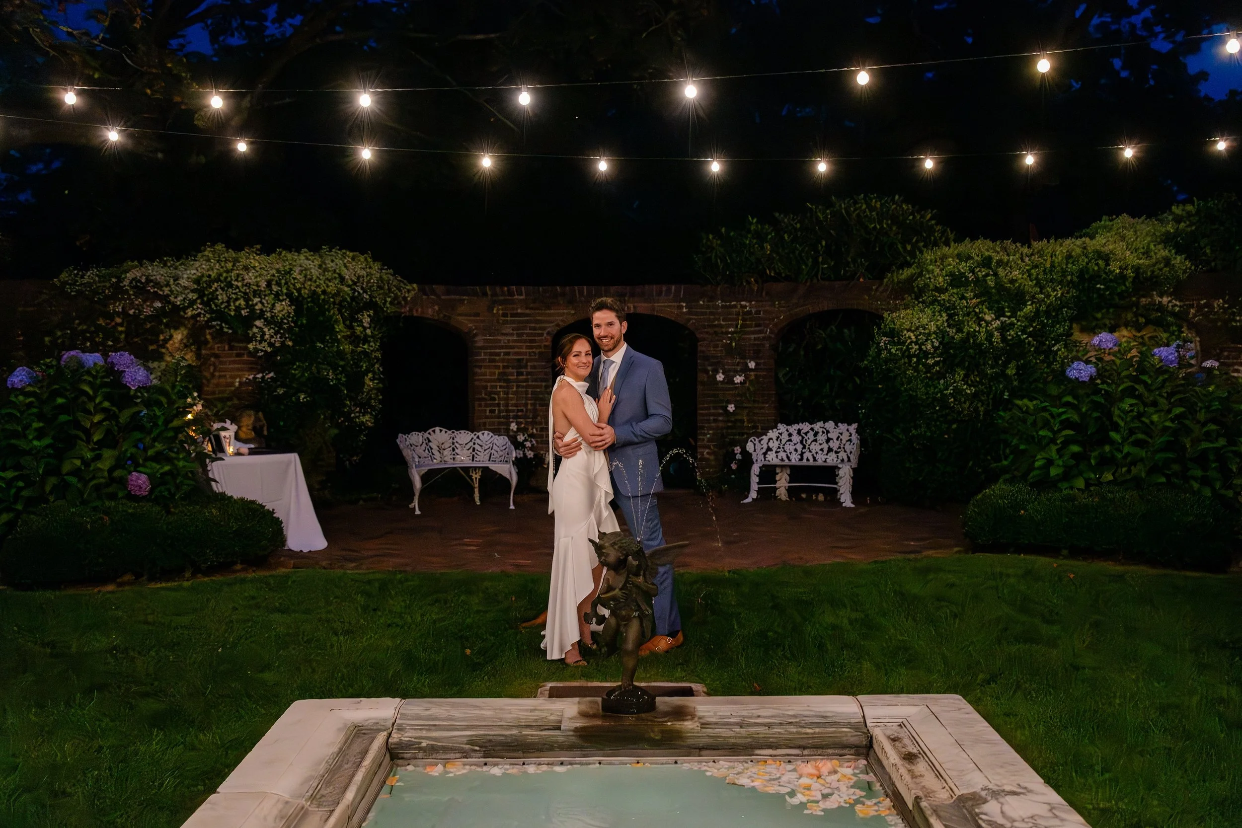 A happy couple dances during their outdoor wedding at night, under hanging string lights, surrounded by lush greenery and flowers, with a decorative fountain in the foreground.