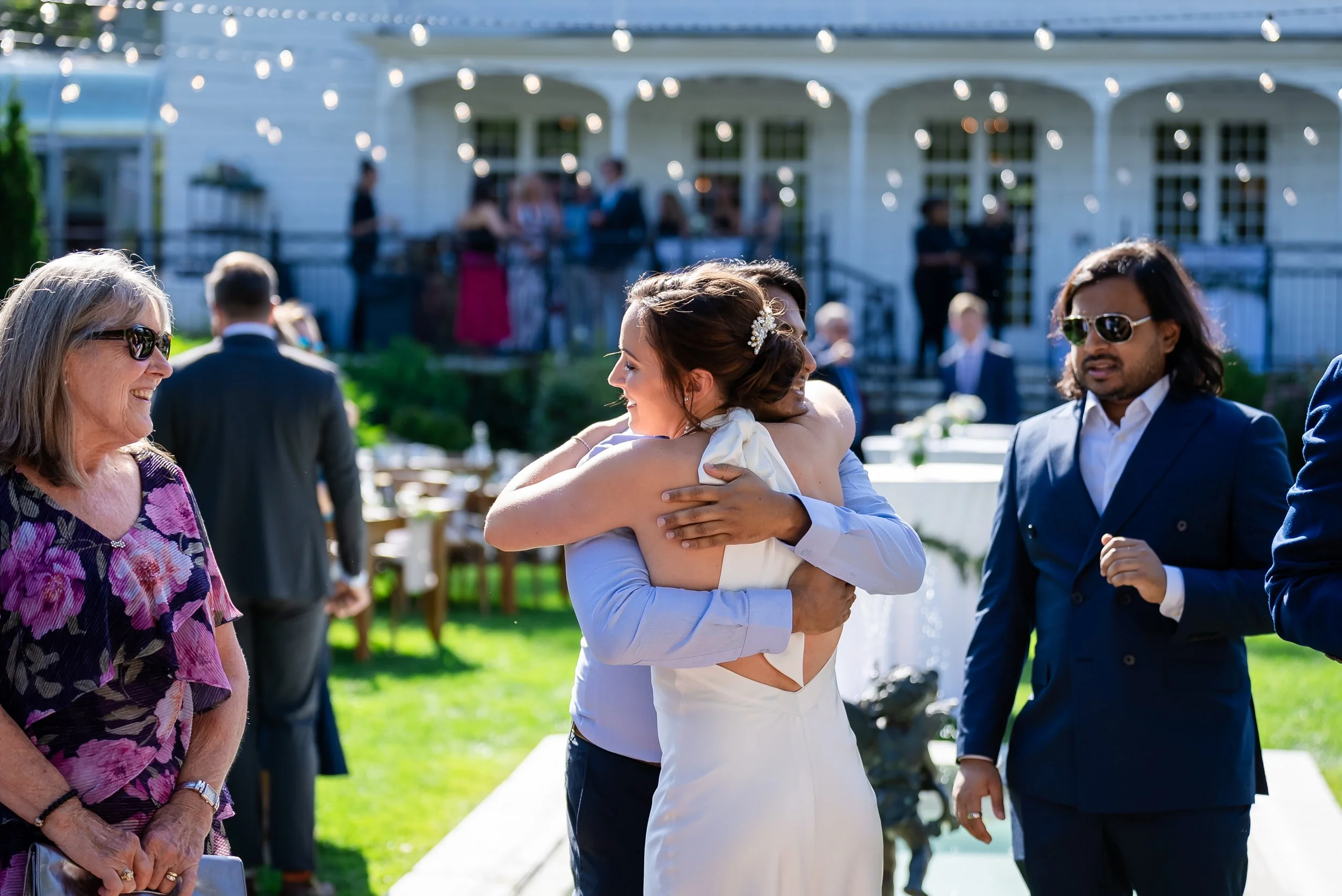 People at a celebration, with two women hugging and others smiling and talking, outdoors in front of a white house with string lights and trees.