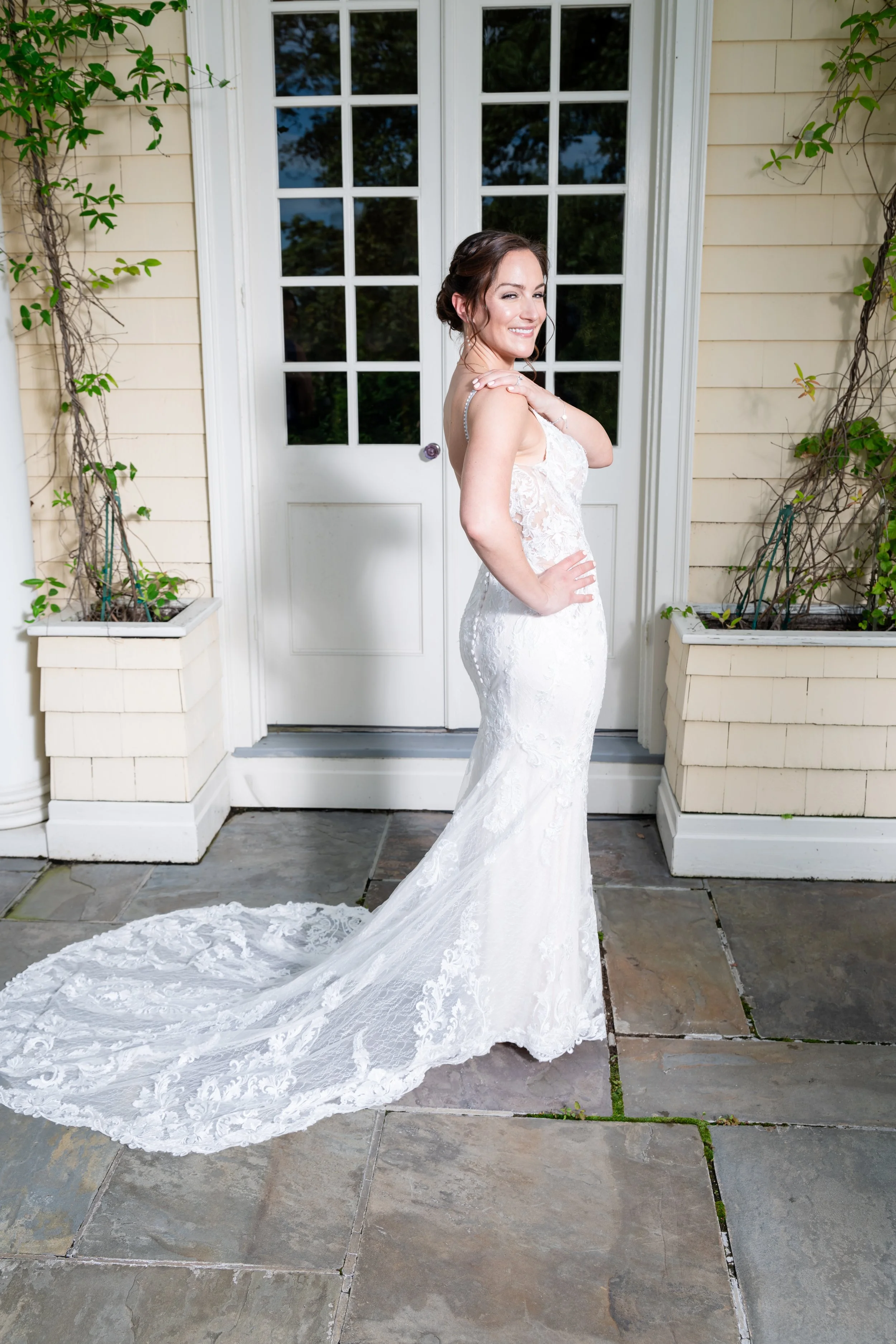 A woman in a white lace wedding dress with a long train stands smiling outside in front of a glass door with white framing, surrounded by beige siding and potted plants with green vines.