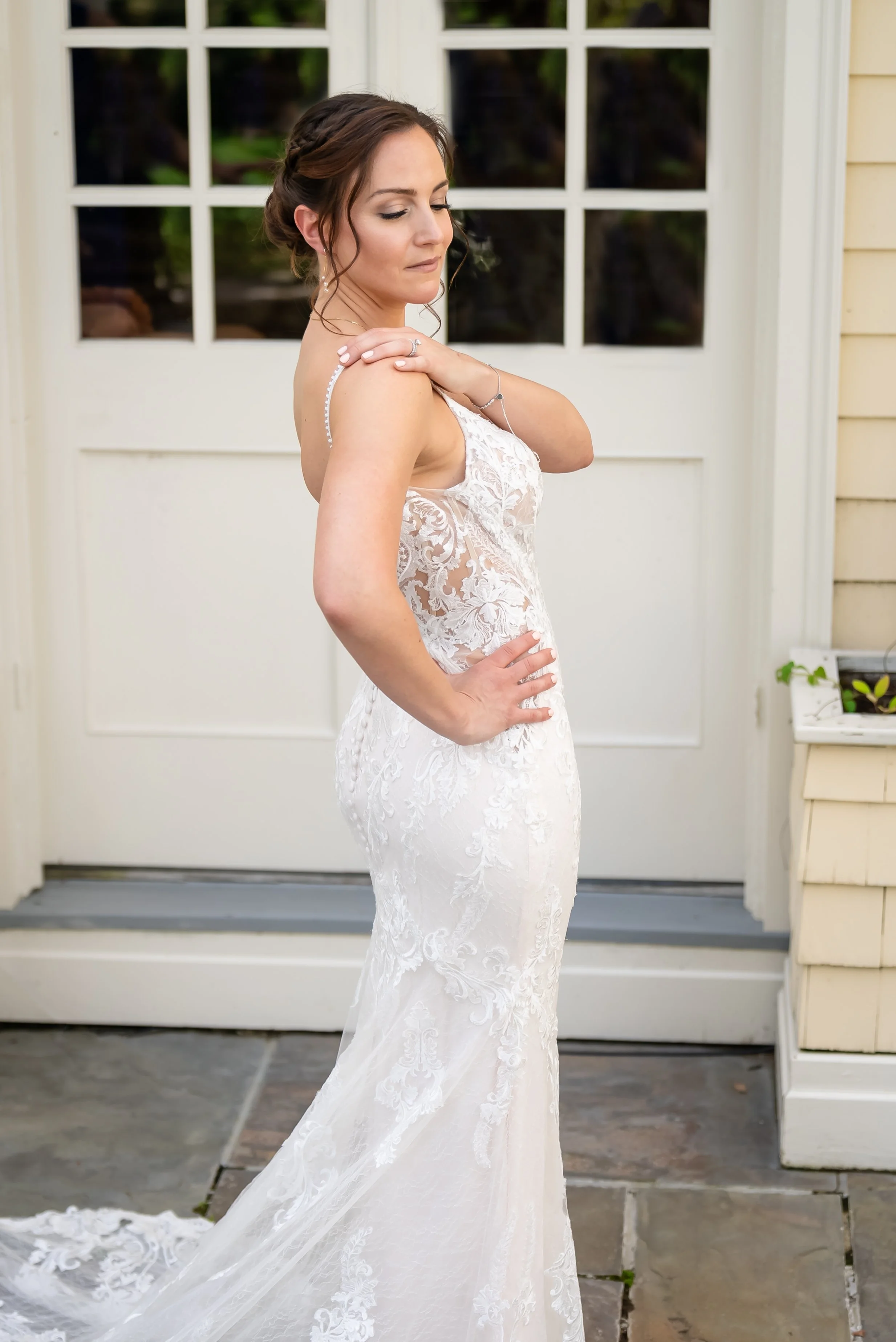 A woman in a white lace wedding dress standing outside in front of a white door with glass panels. She has brown hair styled in an updo and is touching her shoulder with her eyes closed.