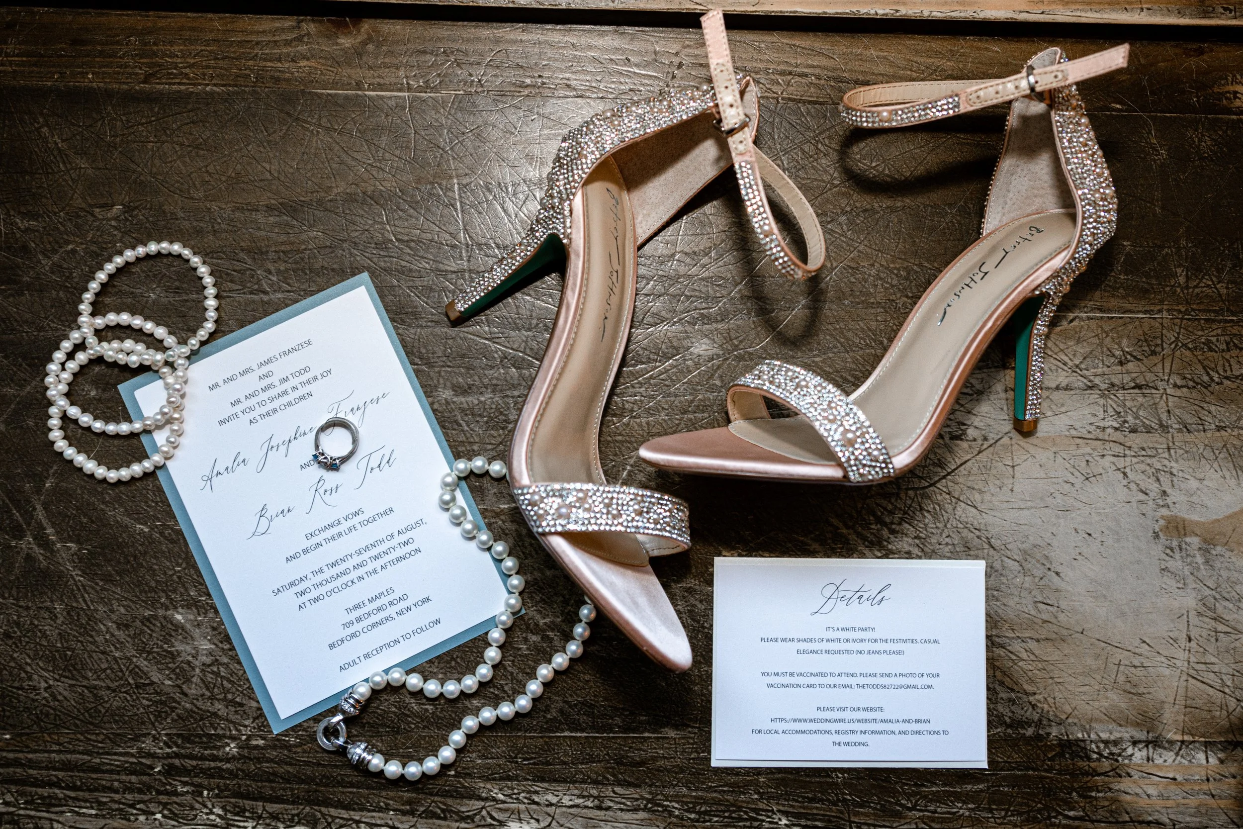 A pair of pink high-heeled shoes decorated with rhinestones, a pearl necklace, and an engagement ring on wedding invitations on a wooden surface.