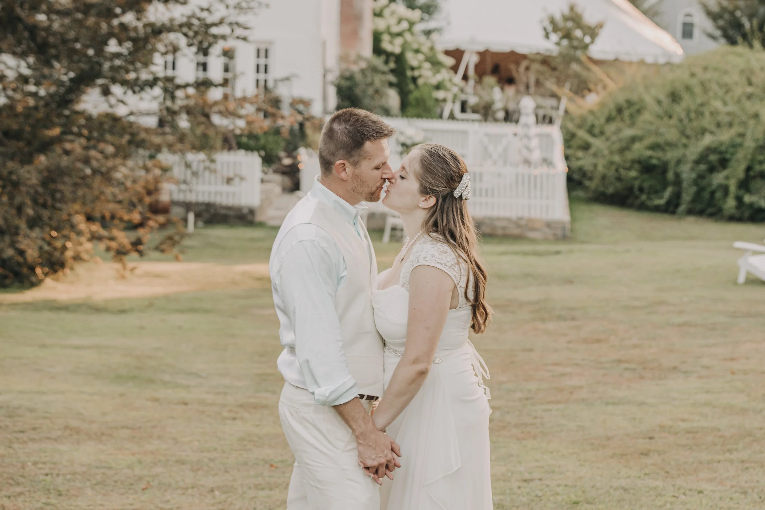 A couple in wedding attire sharing a kiss and holding hands outdoors in a garden setting with a white house in the background.