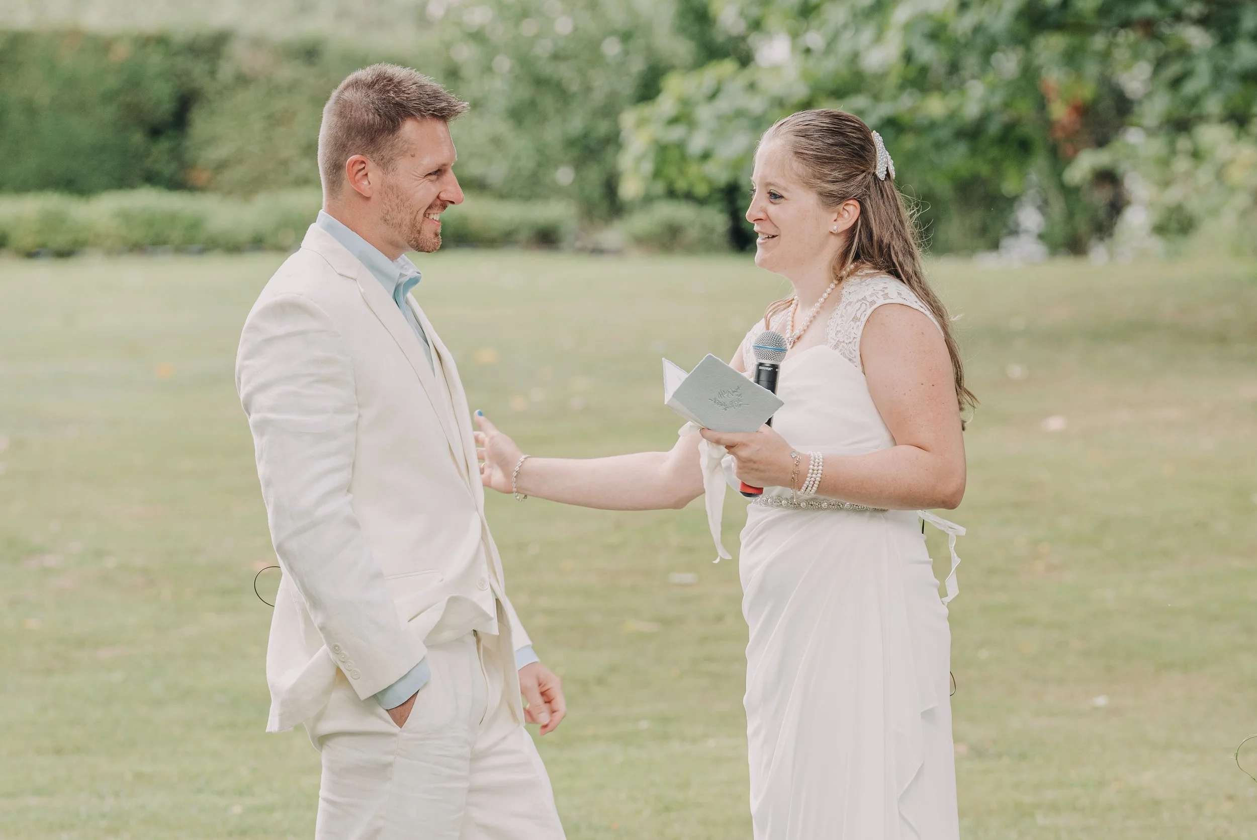 A man and woman during a wedding ceremony outdoors, facing each other and smiling, with the woman holding a microphone and a piece of paper, surrounded by greenery.