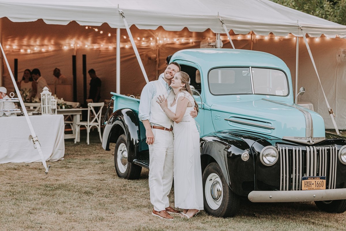 A couple hugging and smiling next to a vintage blue and black truck at a wedding reception under a large tent with string lights and decorated tables.