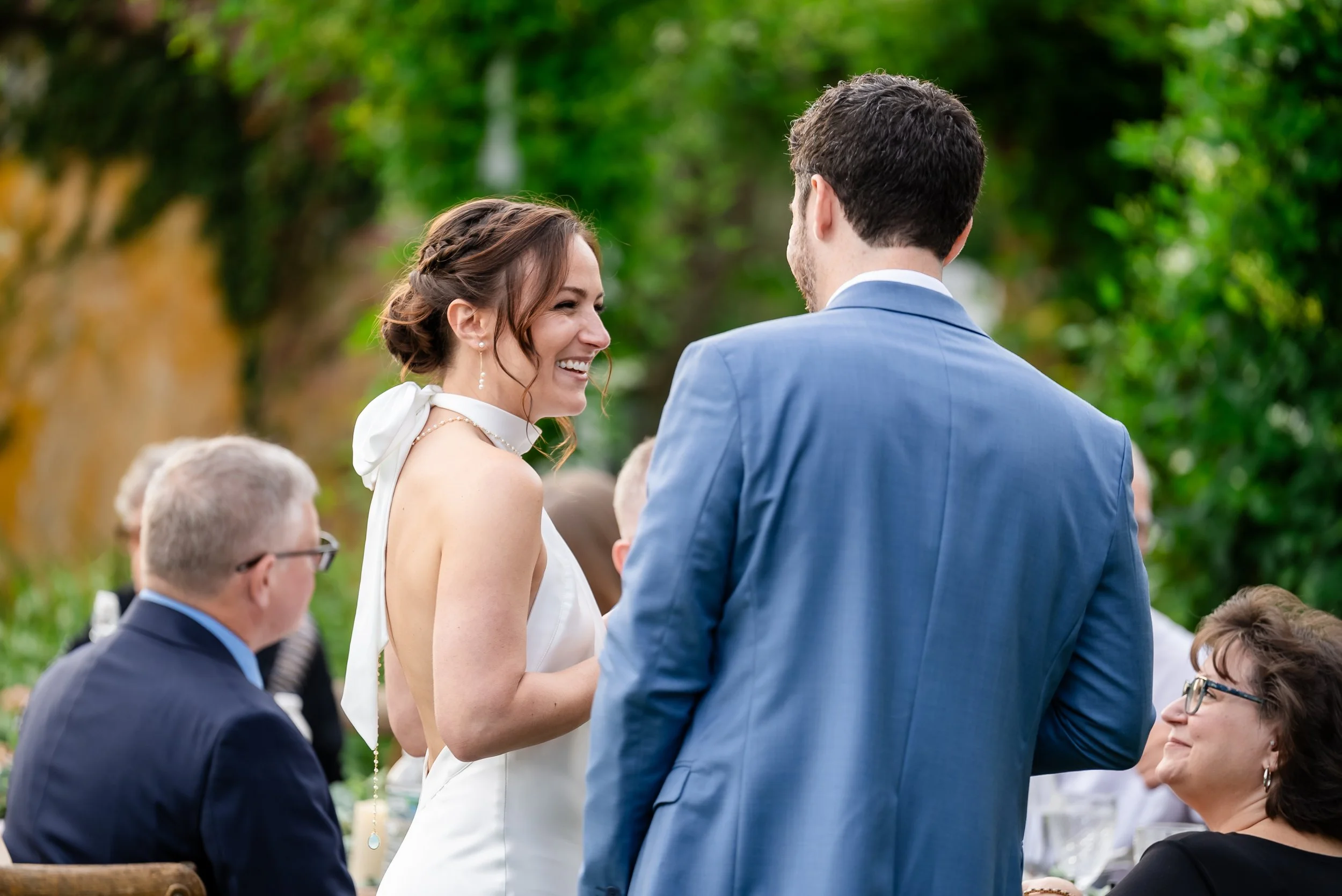 A bride and groom exchanging vows during an outdoor wedding ceremony, with guests seated around them. The bride is smiling and wearing a white dress, while the groom, dressed in a blue suit, is facing her. The setting is lush with green foliage in th