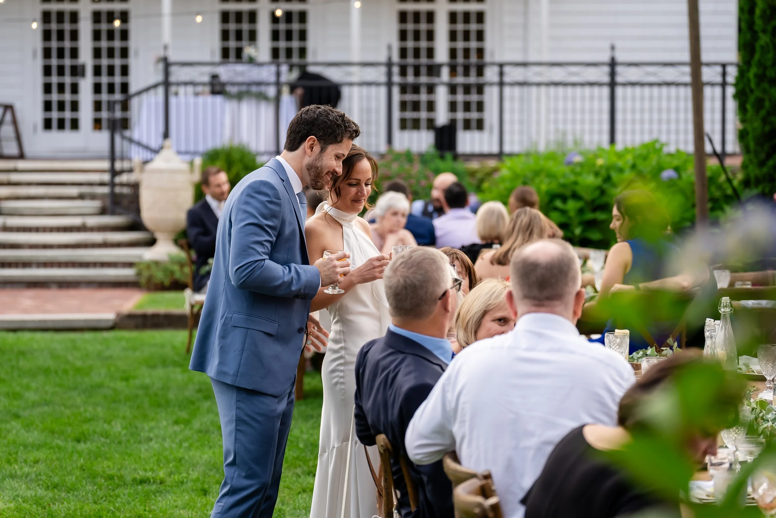 A couple, dressed in formal wedding attire, standing outdoors during a wedding reception, holding glasses and smiling amongst seated guests at a garden party.
