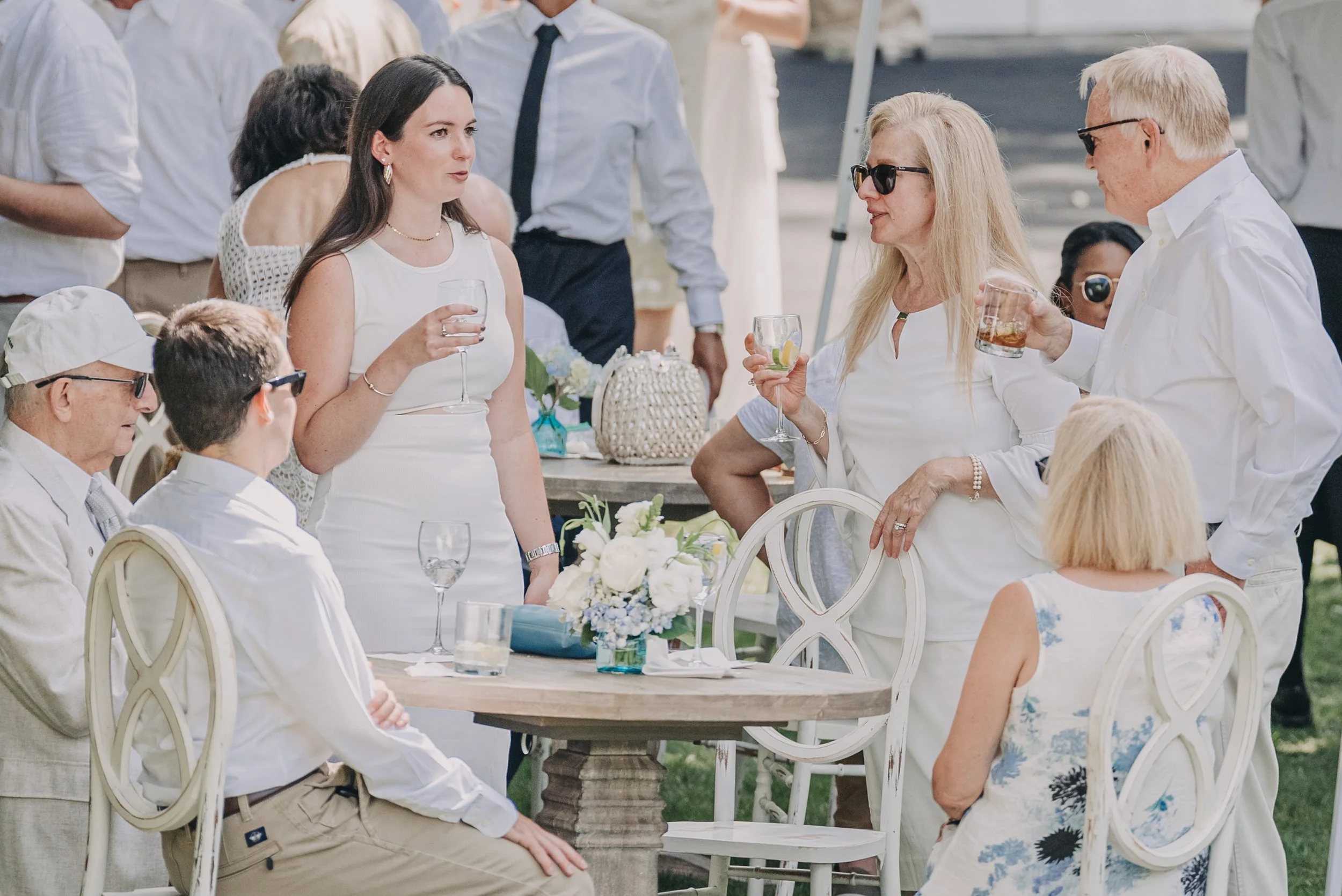 People gathered at a white outdoor event table, socializing with drinks, floral centerpiece, and white chairs, during daytime.