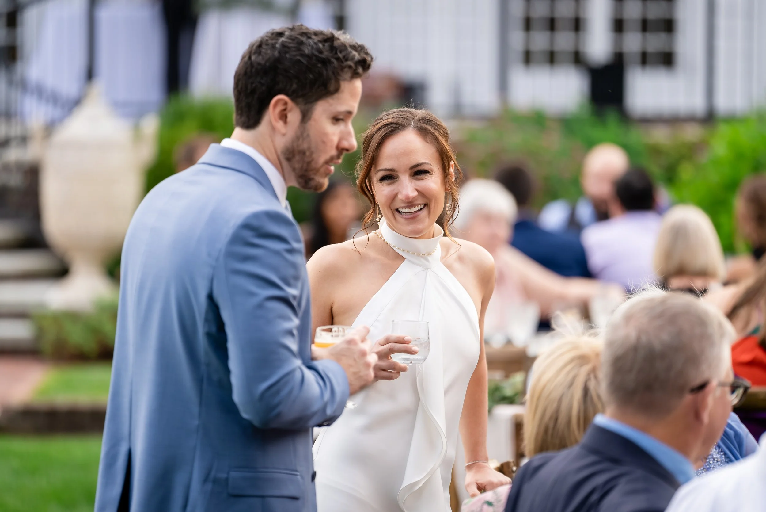 A man in a blue suit and a woman in a white dress holding wine glasses at an outdoor social event with several people seated in the background.