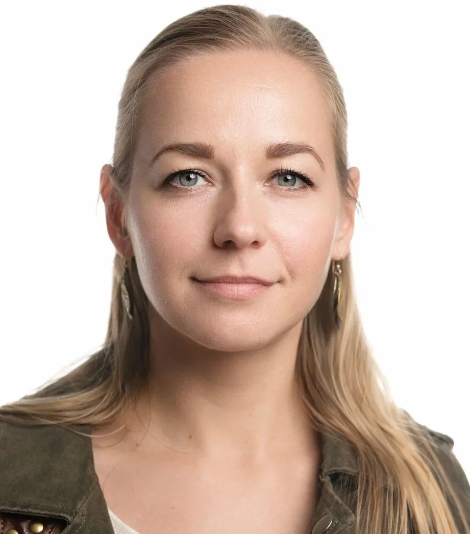 Close-up portrait of a woman with long blonde hair, blue eyes, and fair skin, smiling softly, wearing earrings and a green military-style jacket against a white background.