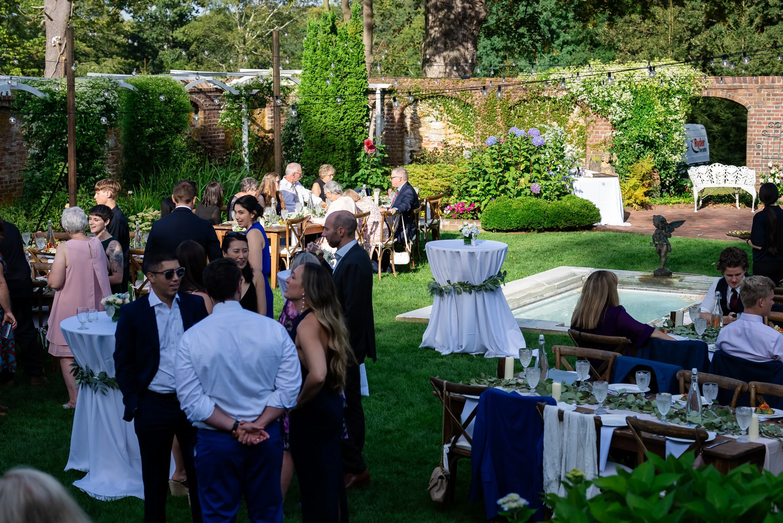 An outdoor celebration or wedding reception with guests seated at decorated tables, lush greenery, and string lights hanging between trees and brick walls.