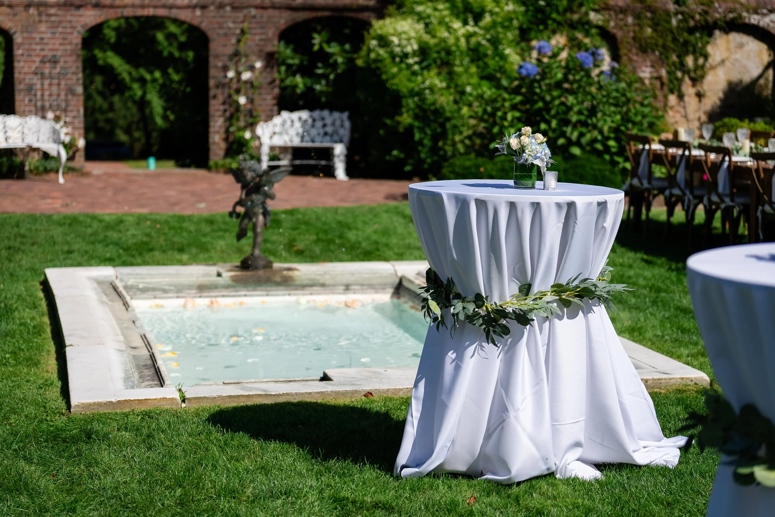Outdoor wedding reception setup with a round table covered with a white draped cloth, decorated with a floral centerpiece in a green vase, set in a garden with chairs and greenery in the background.