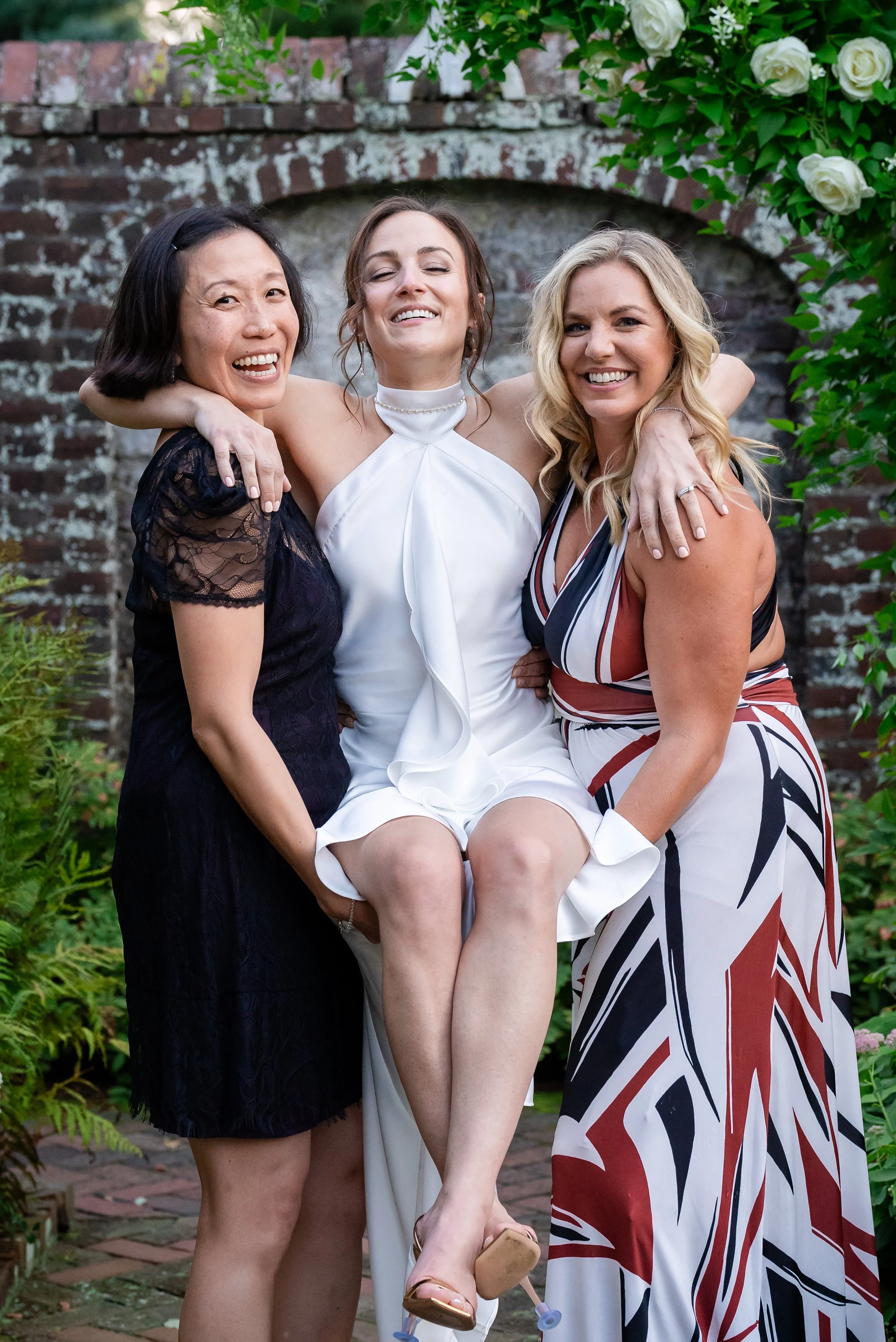 Three women smiling and hugging each other outdoors in front of a brick wall with white roses and green foliage. The woman in the middle is wearing a white dress, and the women on either side are dressed in black and multicolored dresses. The woman o