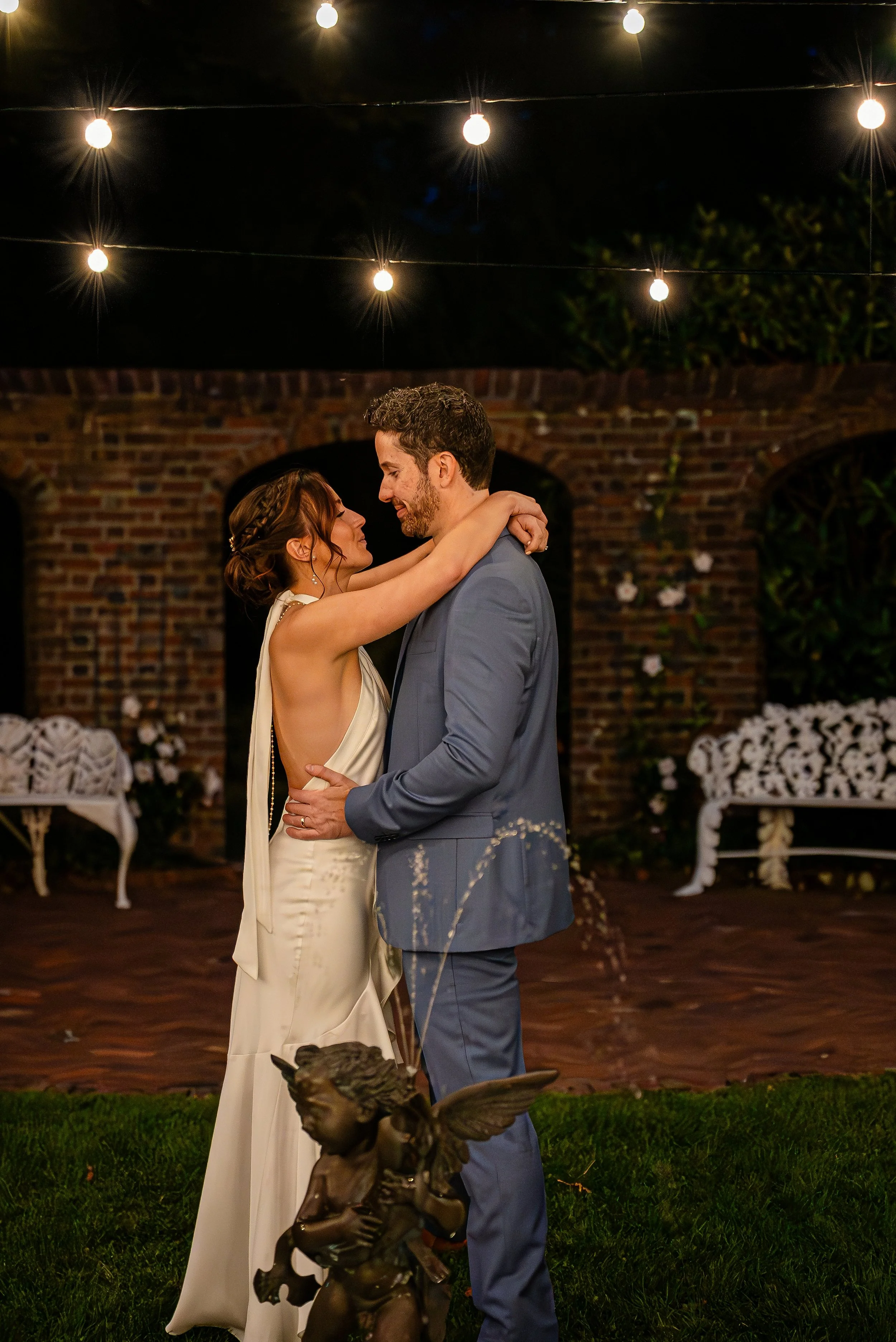A bride and groom are dancing at their wedding reception under string lights, with brick walls and floral decorations in the background.