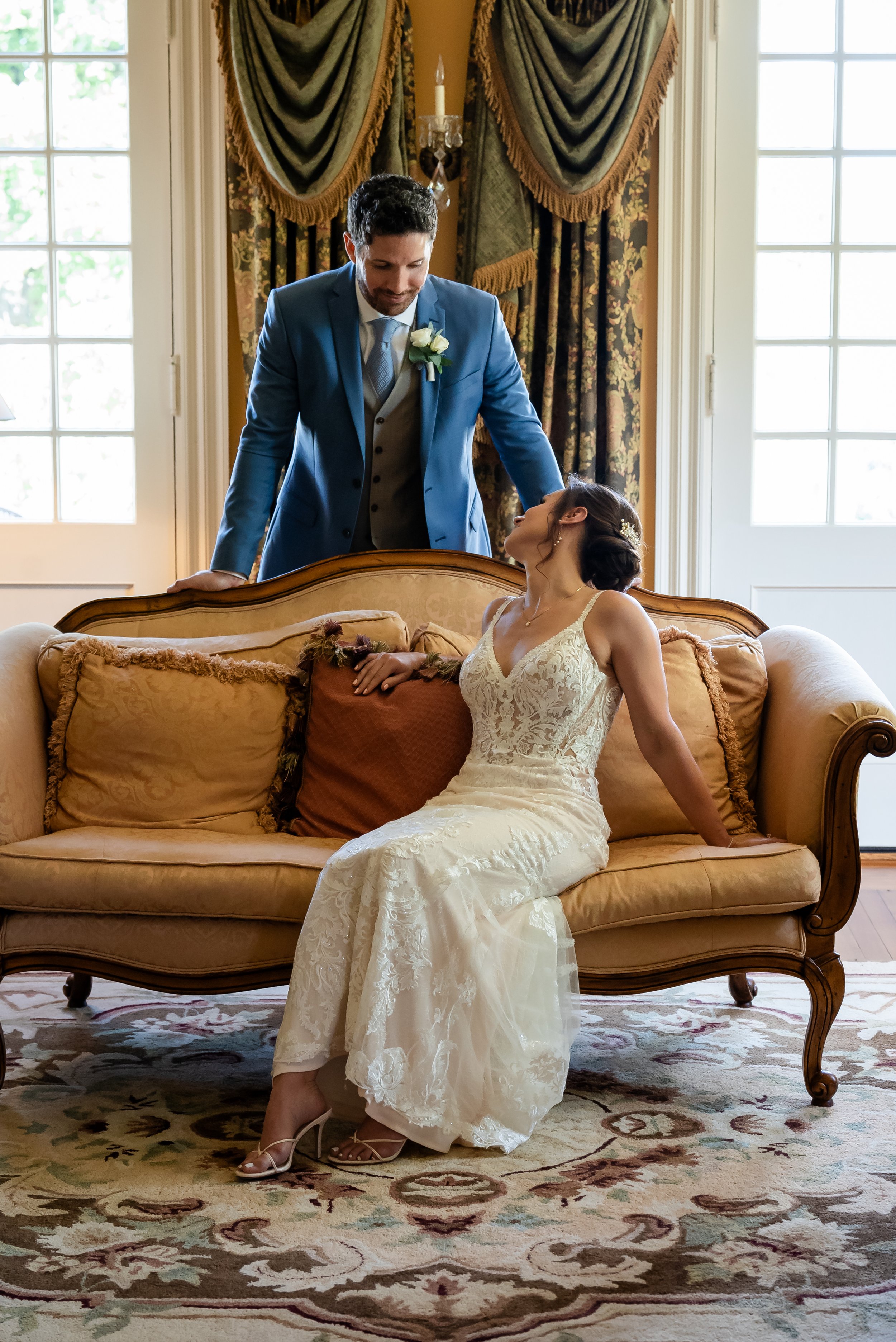 A bride in a white lace wedding dress sitting on a vintage sofa, looking up at a groom in a blue suit who is leaning over the sofa. The scene takes place in an elegantly decorated room with large windows and dark drapes.