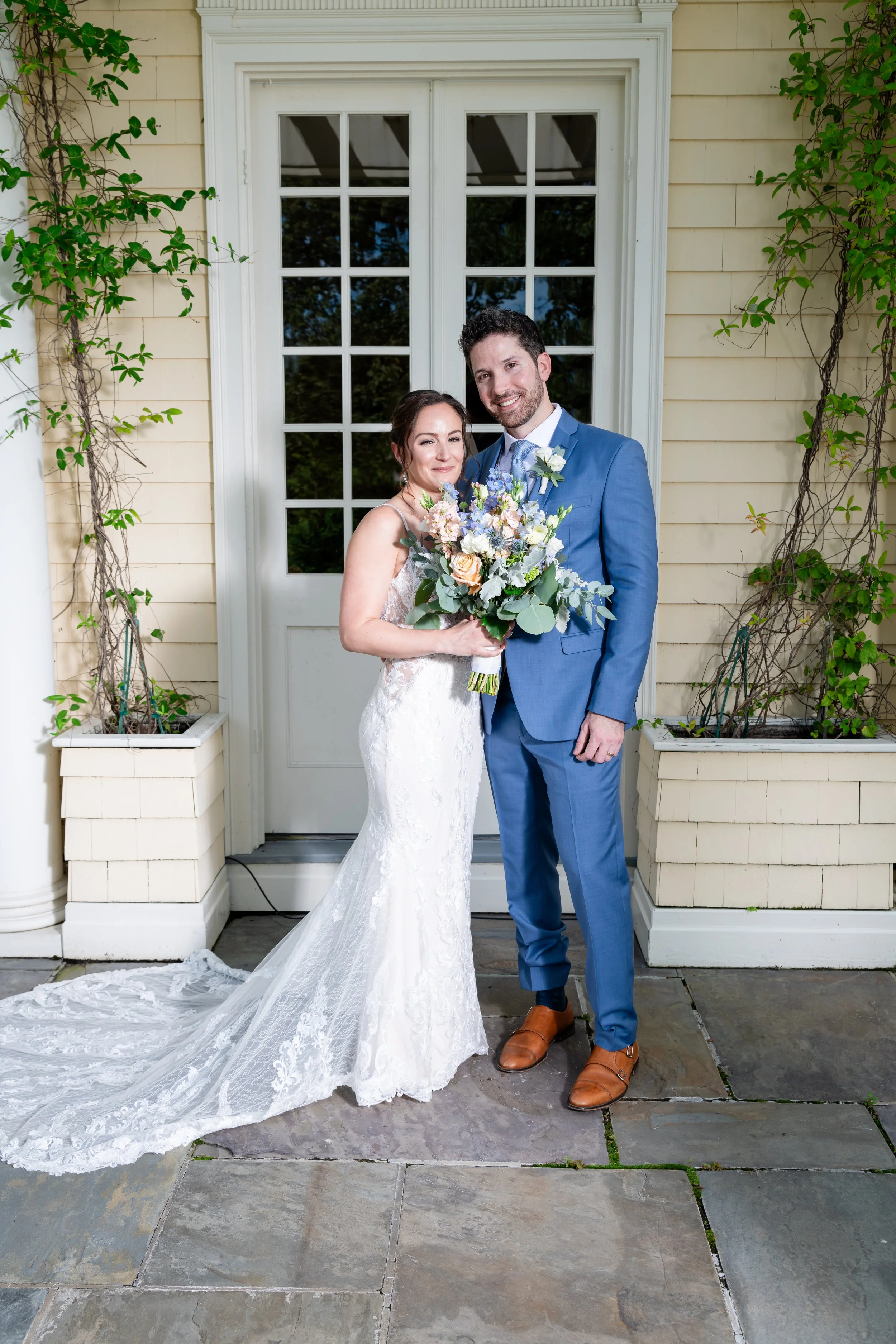 A bride and groom stand close together outdoors, smiling at the camera. The bride wears a lace wedding gown with a long train and holds a bouquet of flowers. The groom wears a blue suit with a light blue shirt, a boutonniere, and brown shoes. They ar